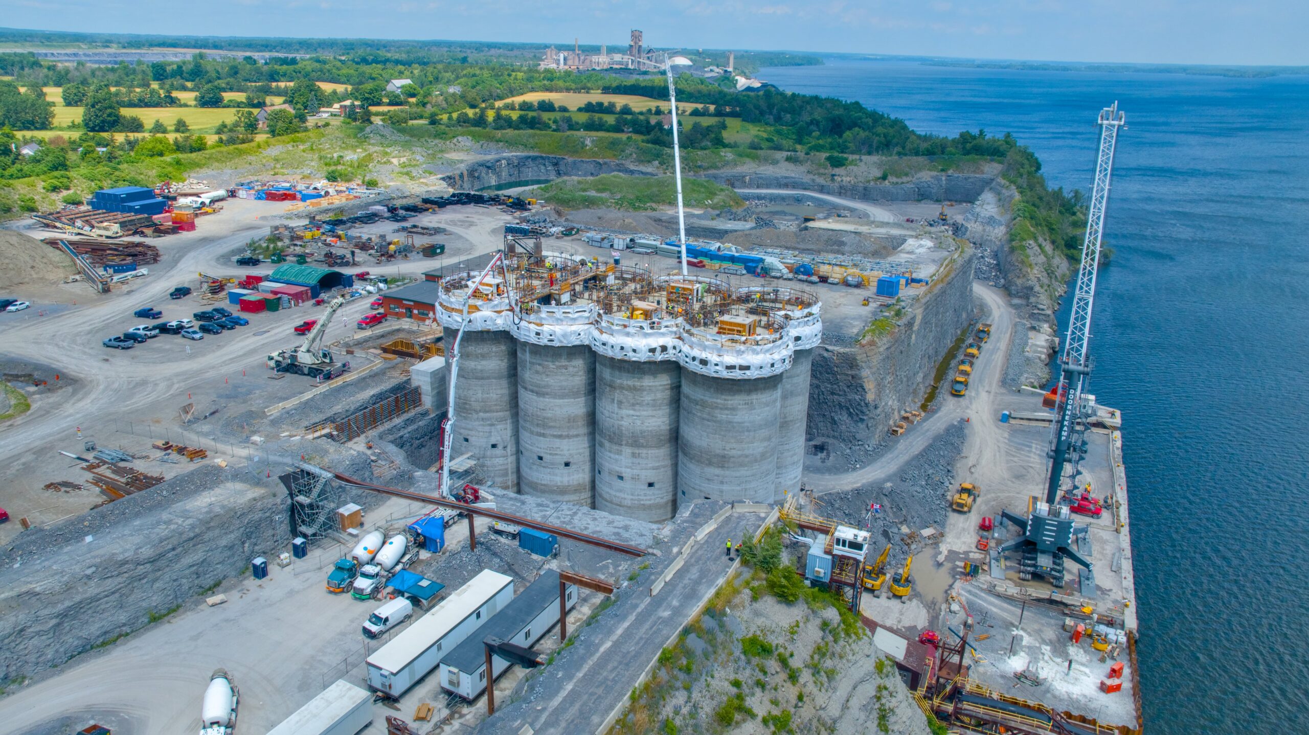 Aerial view of a waterfront construction site with multiple large cylindrical silos, cranes, machinery, and nearby water.