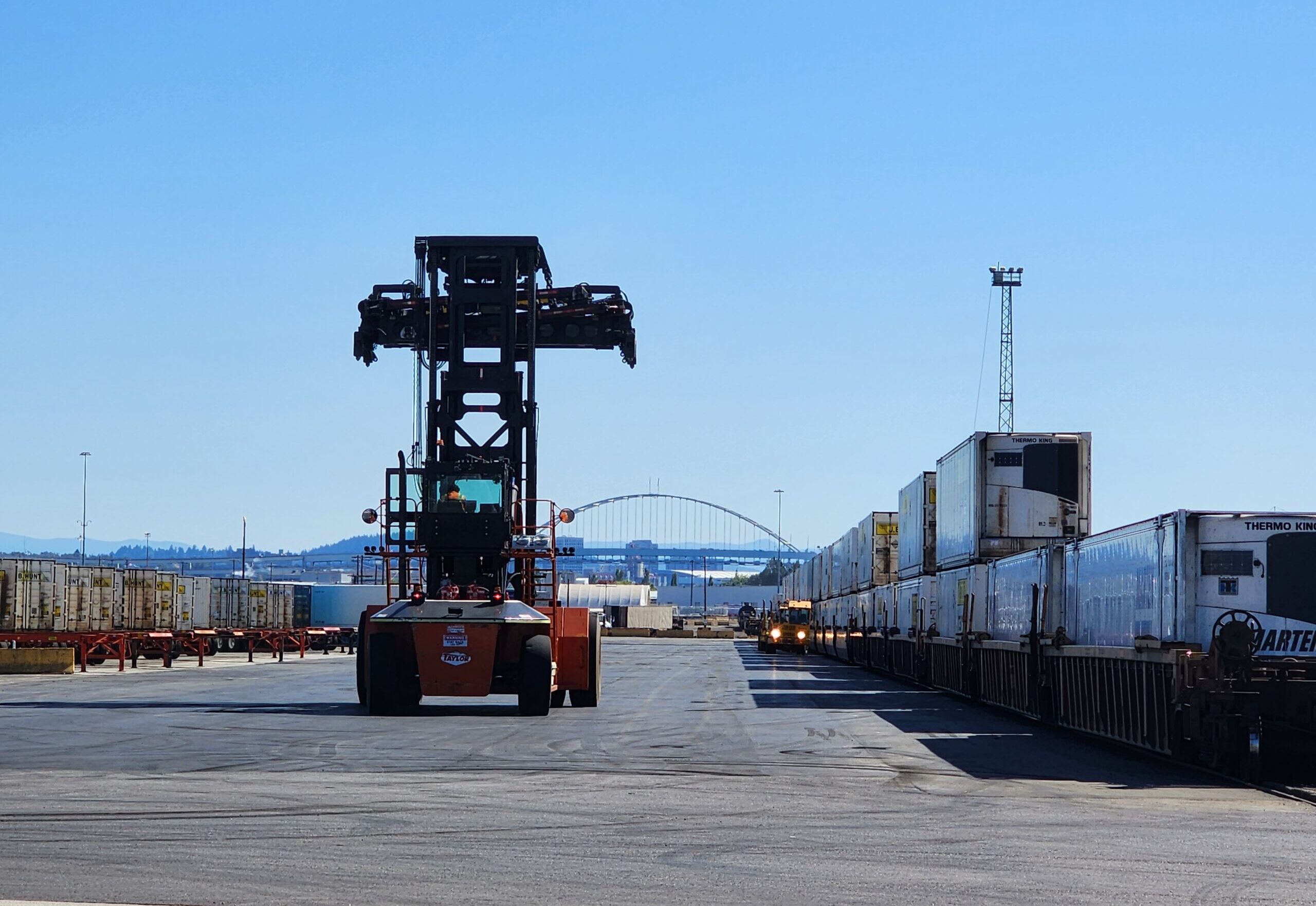A large container handler operates in a rail yard lined with freight trains under a clear blue sky, with a bridge visible in the background.