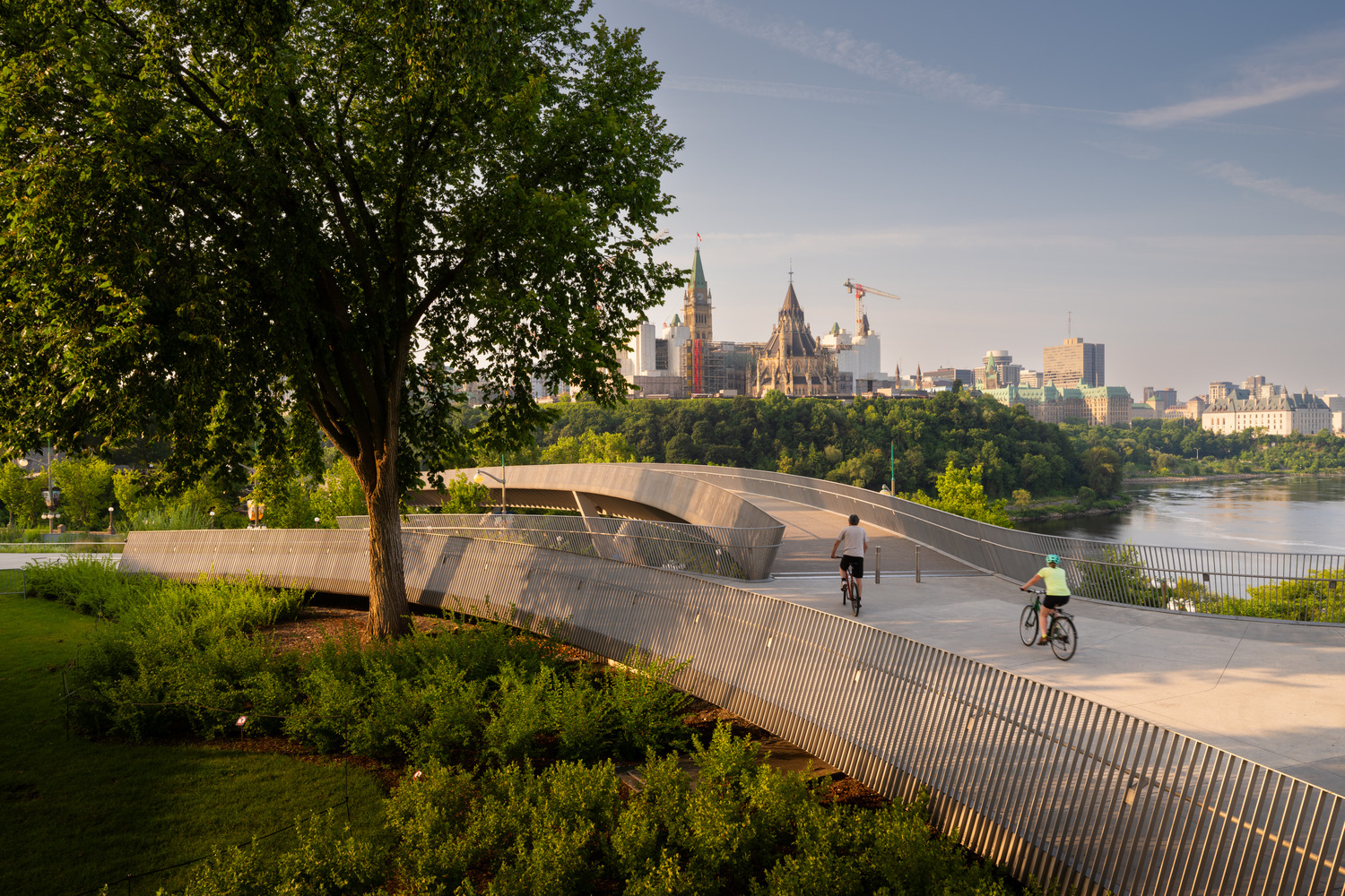 Two cyclists ride on a modern elevated pathway with trees and greenery, with historic parliament buildings and city skyline in the background.