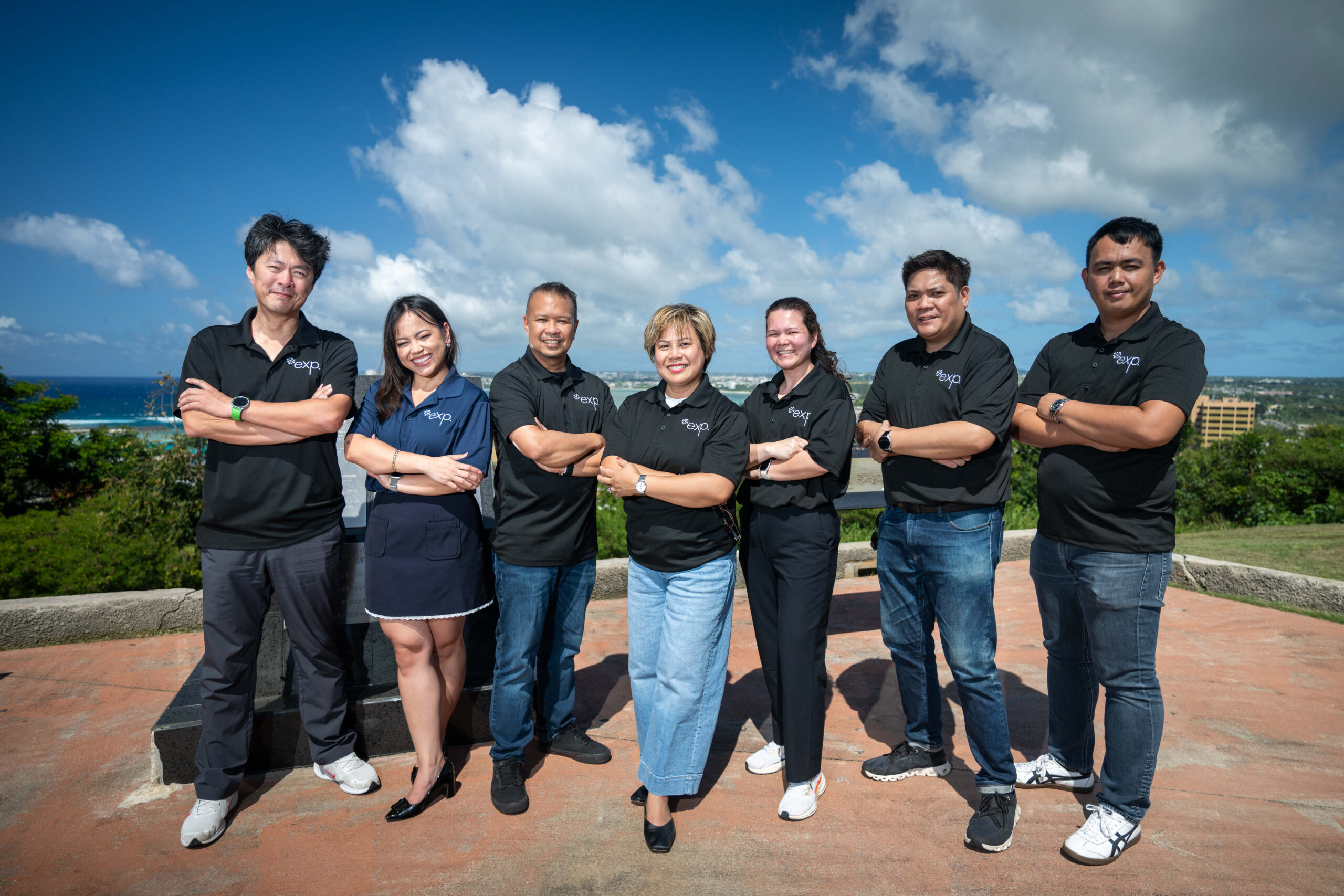 Seven people stand outdoors with arms crossed, smiling at the camera in dark logo shirts, as trees, ocean, and a cloudy sky showcase Guam infrastructure resilience in the background.