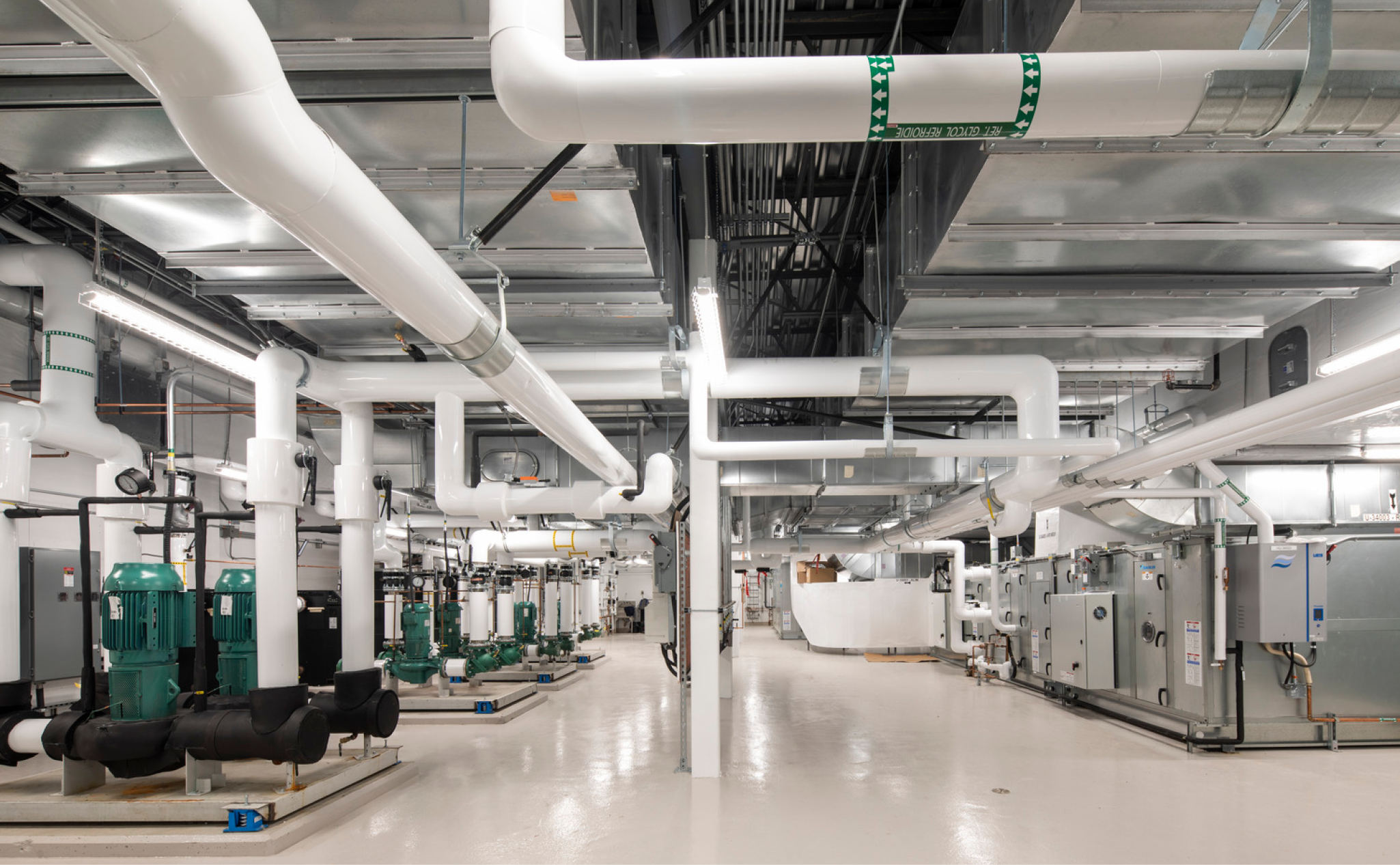 Intérieur d'un local technique avec des tuyaux isolés blancs, des pompes vertes, des unités de traitement de l'air et des tuyaux de glycol étiquetés qui courent le long du plafond.