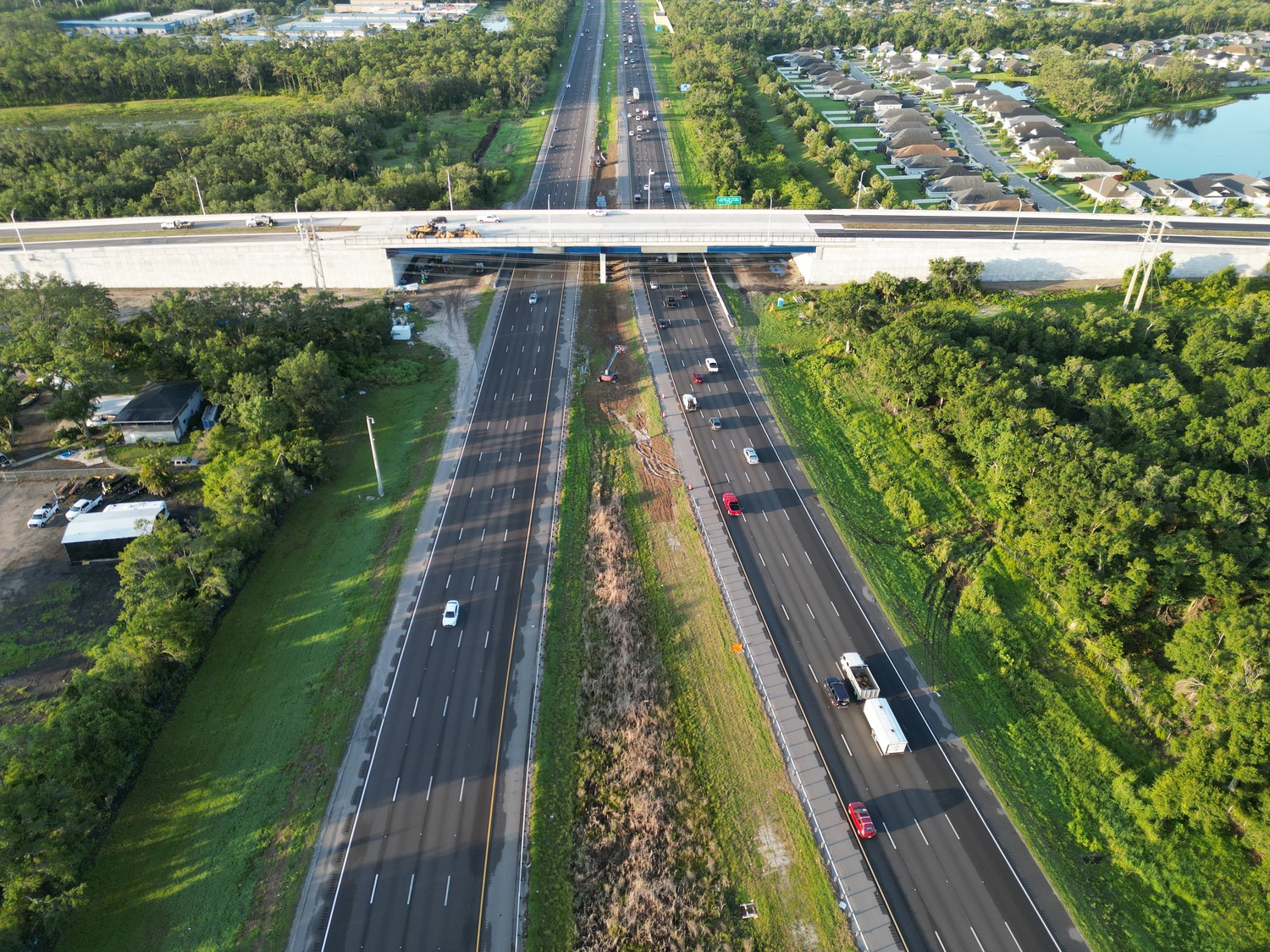 Aerial view of a divided highway with light traffic, an overpass under construction, and nearby residential and wooded areas.