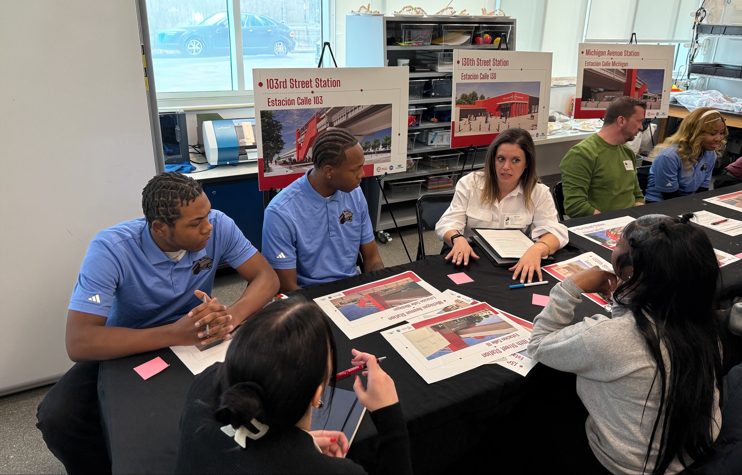 A group of people sit at a table discussing the Red Line Extension station, with posters of the 103rd Street Station displayed in the background.