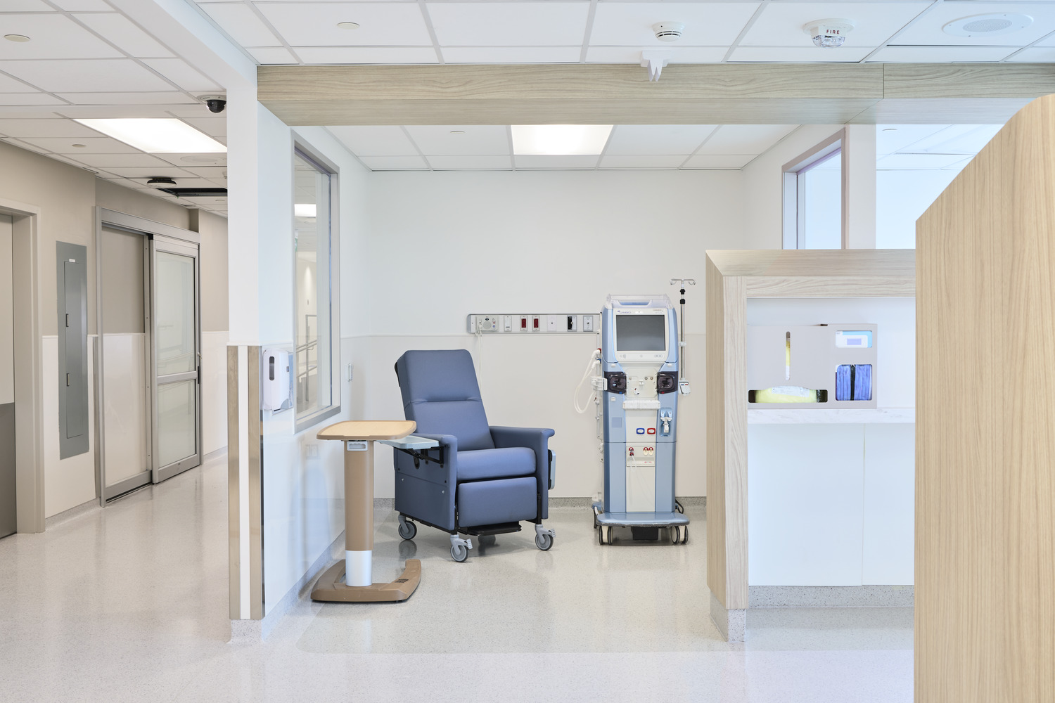 A medical treatment room with a blue reclining chair, medical equipment, and a small table, featuring clean, modern design and bright lighting.