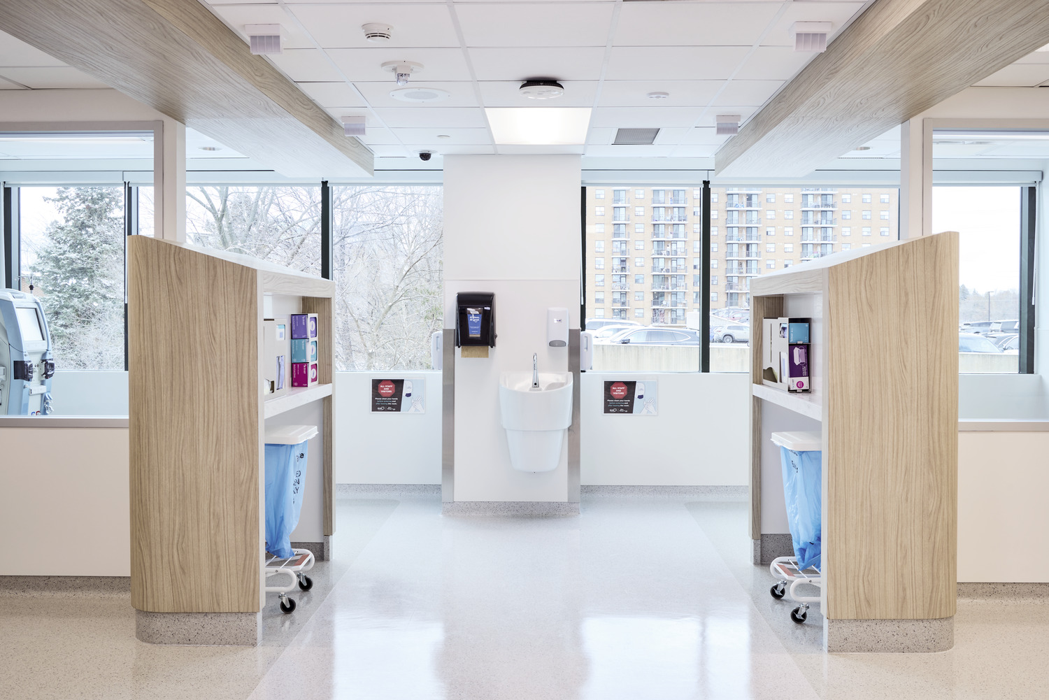 A modern hospital room with two medical workstations facing each other, a central handwashing sink, and large windows showing buildings and trees outside.