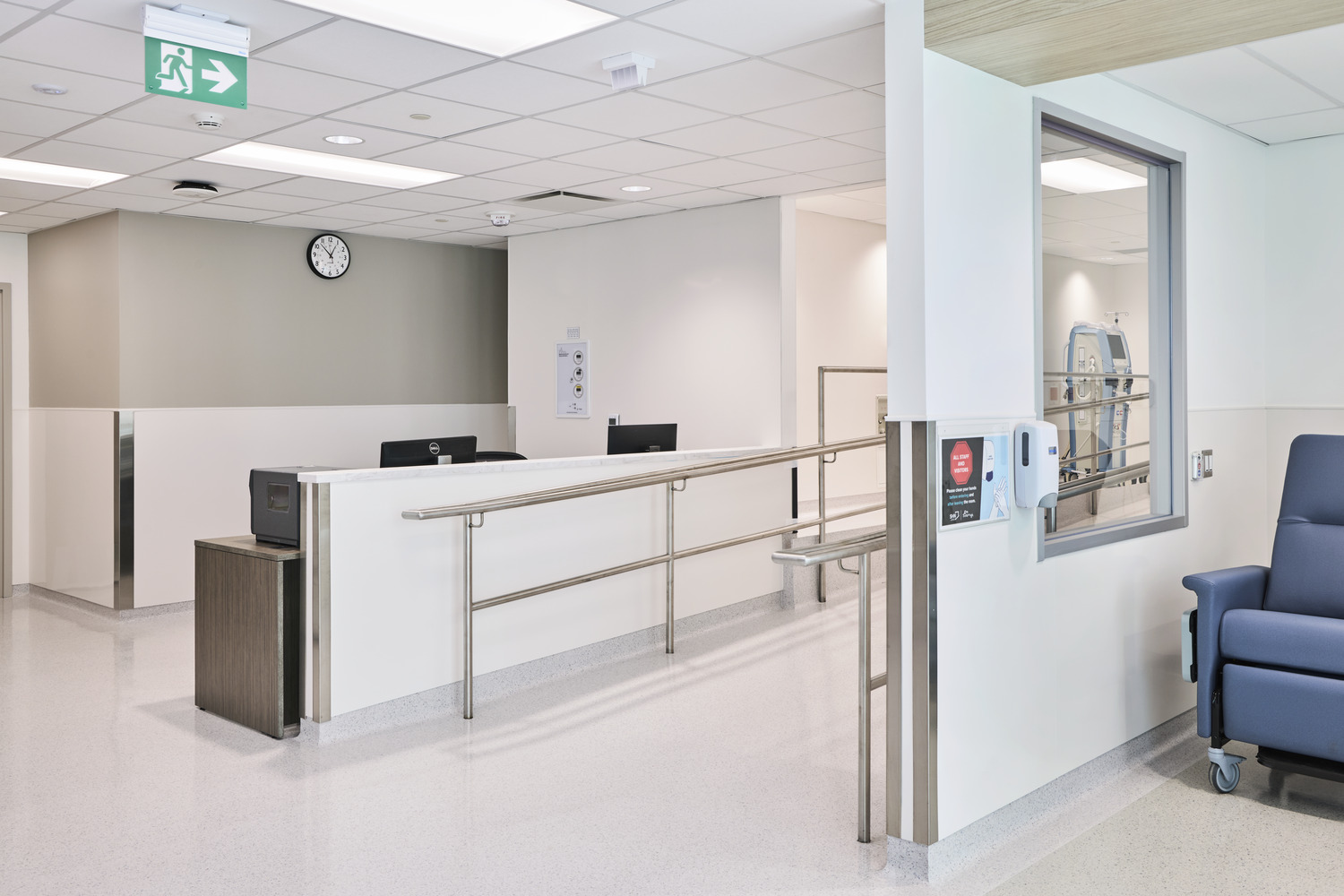 A clean, empty hospital reception area with a desk, computers, safety railings, wall clock, and an empty blue recliner near a window.