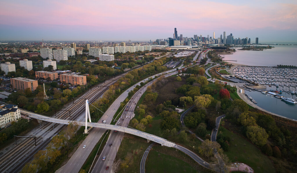 Aerial view of a city blending transportation engineering features like a highway, pedestrian bridge, residential buildings, marina, and a distant downtown skyline at sunset.