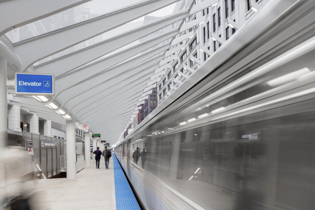 A train moves through a modern, bright subway station with people waiting on the platform and an elevator sign visible, showcasing the impact of transportation engineering.