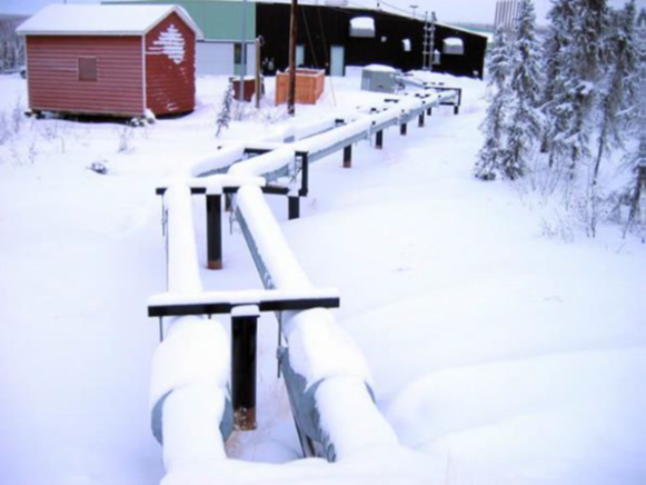 Snow-covered pipelines run above ground near small buildings and trees in a winter landscape.