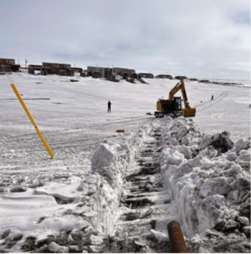 A yellow excavator clears snow on a wide, snowy landscape with buildings in the background and a yellow pole leaning to the left in the foreground.