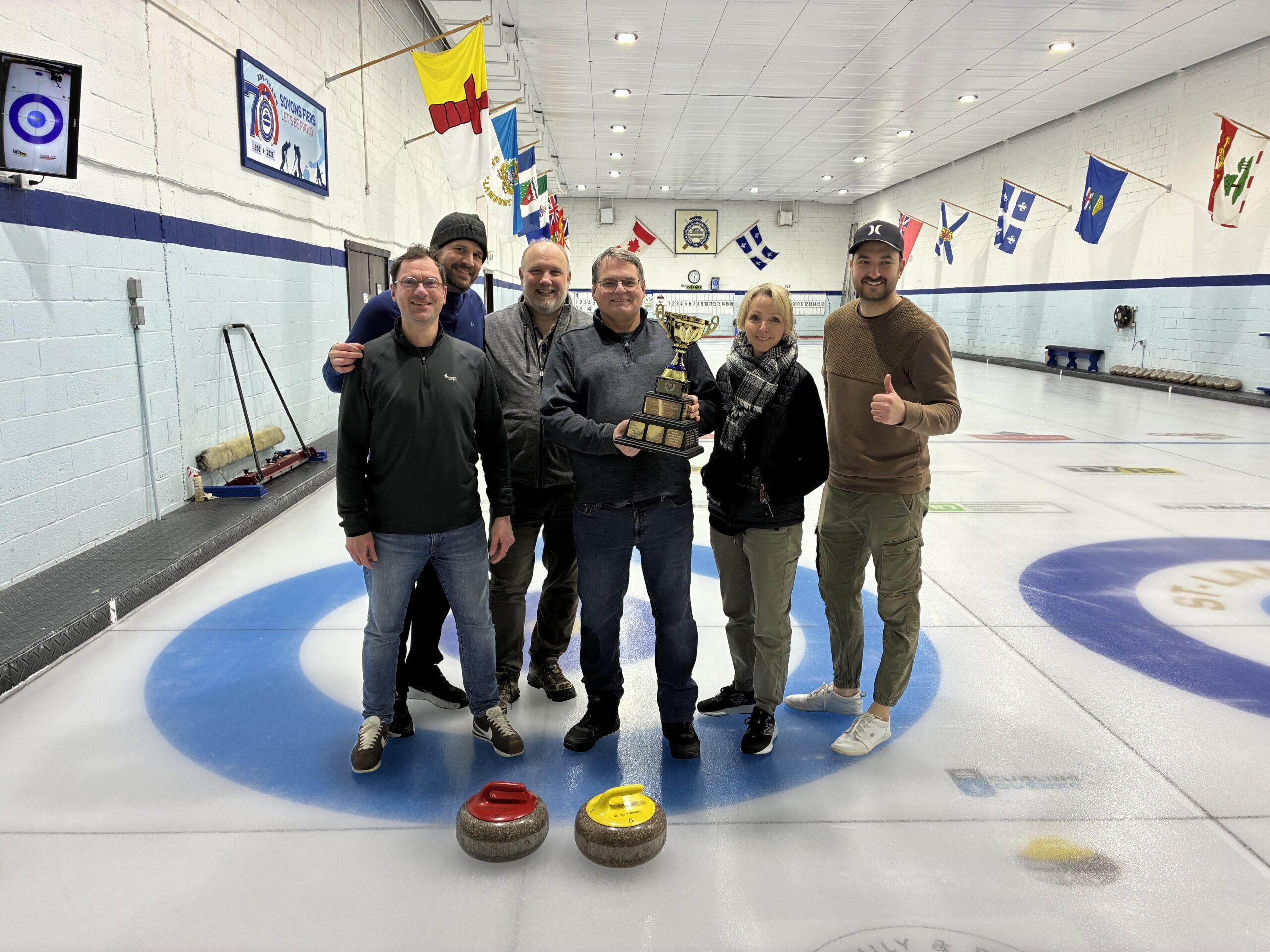 Six personnes se tiennent sur une piste de curling, l'une d'entre elles tenant un trophée. Deux pierres de curling sont posées sur la glace devant eux et des drapeaux nationaux sont suspendus au-dessus d'eux.