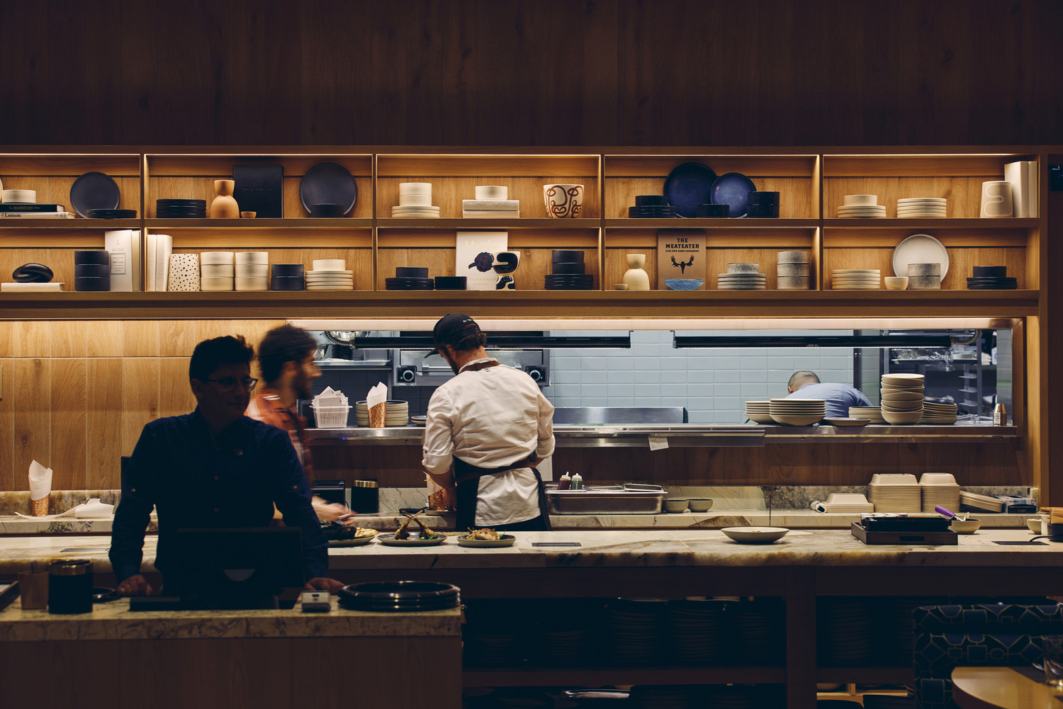 Two chefs work in an all-electric restaurant kitchen behind a counter, preparing food, while a person stands in the dimly lit foreground. Shelves with plates and dishes are visible on the wall at Limelight Boulder.