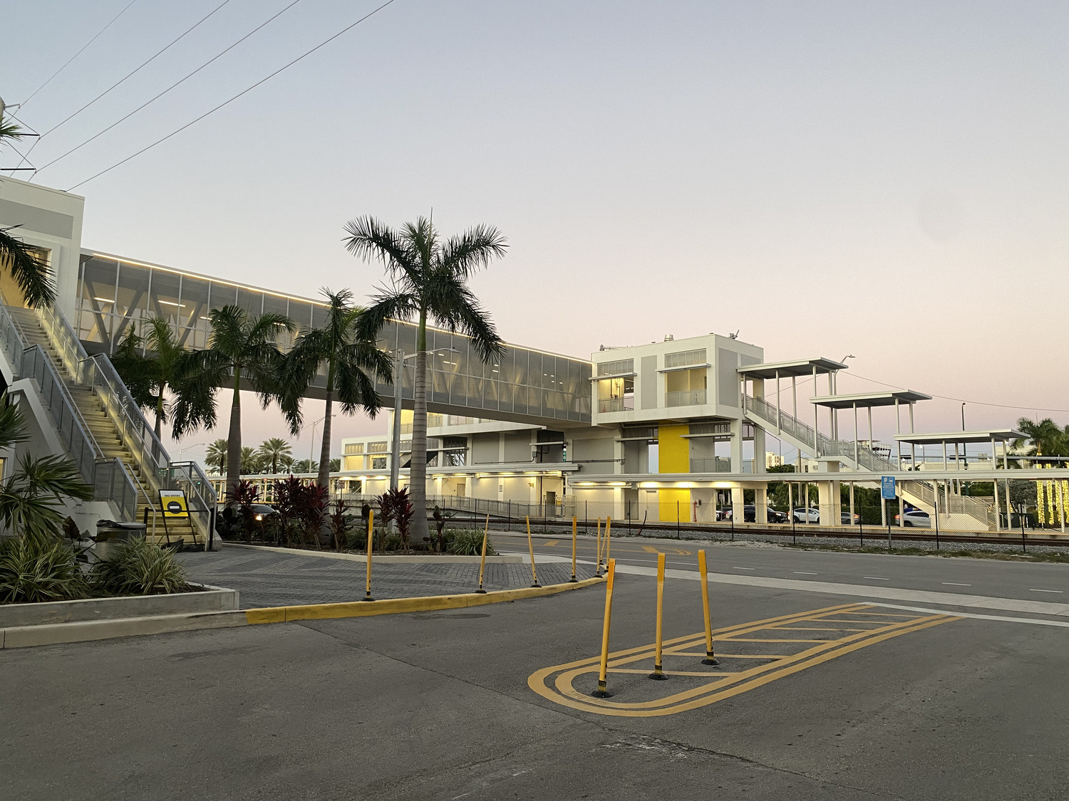 Modern building complex with a glass walkway, palm trees, and a small parking area at sunset. Yellow accents highlight the structure.