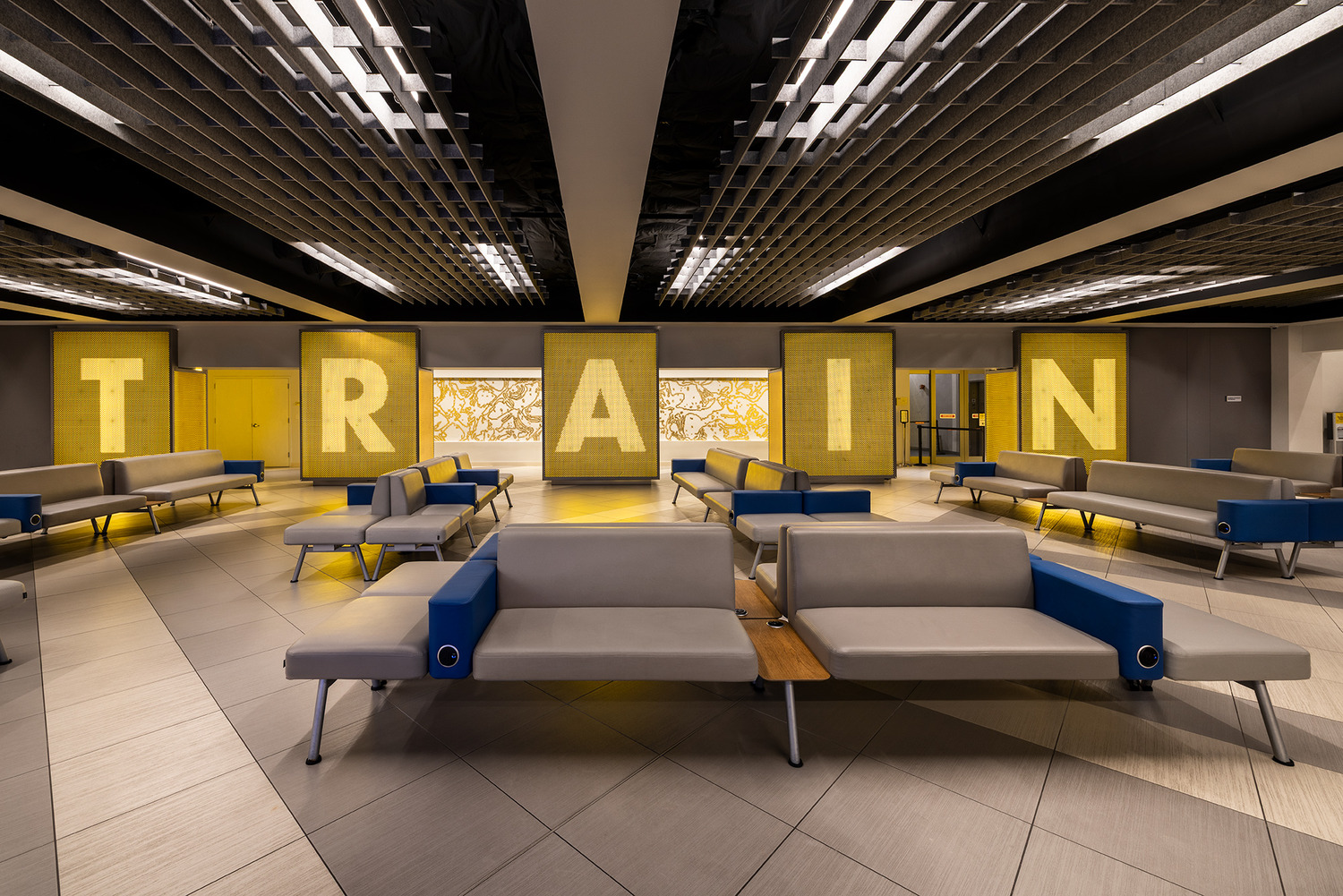 Modern waiting area with gray and blue benches; large illuminated letters on the back wall spell 