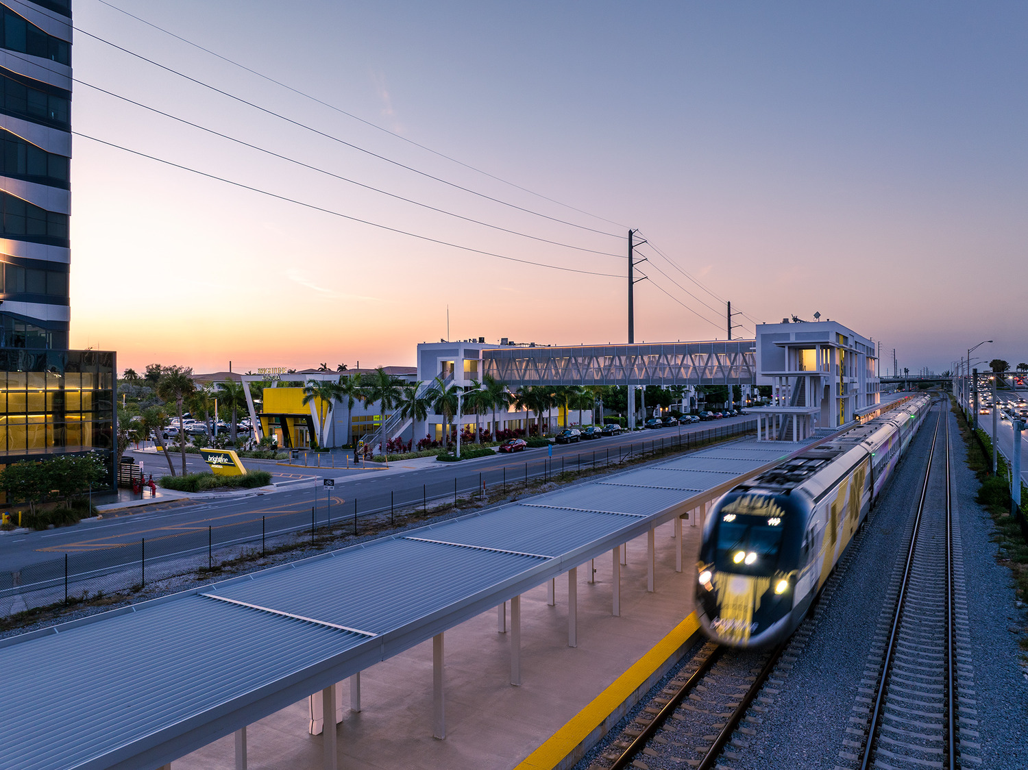 A passenger train arrives at a modern outdoor station at sunset, with palm trees, a pedestrian overpass, and surrounding buildings visible.