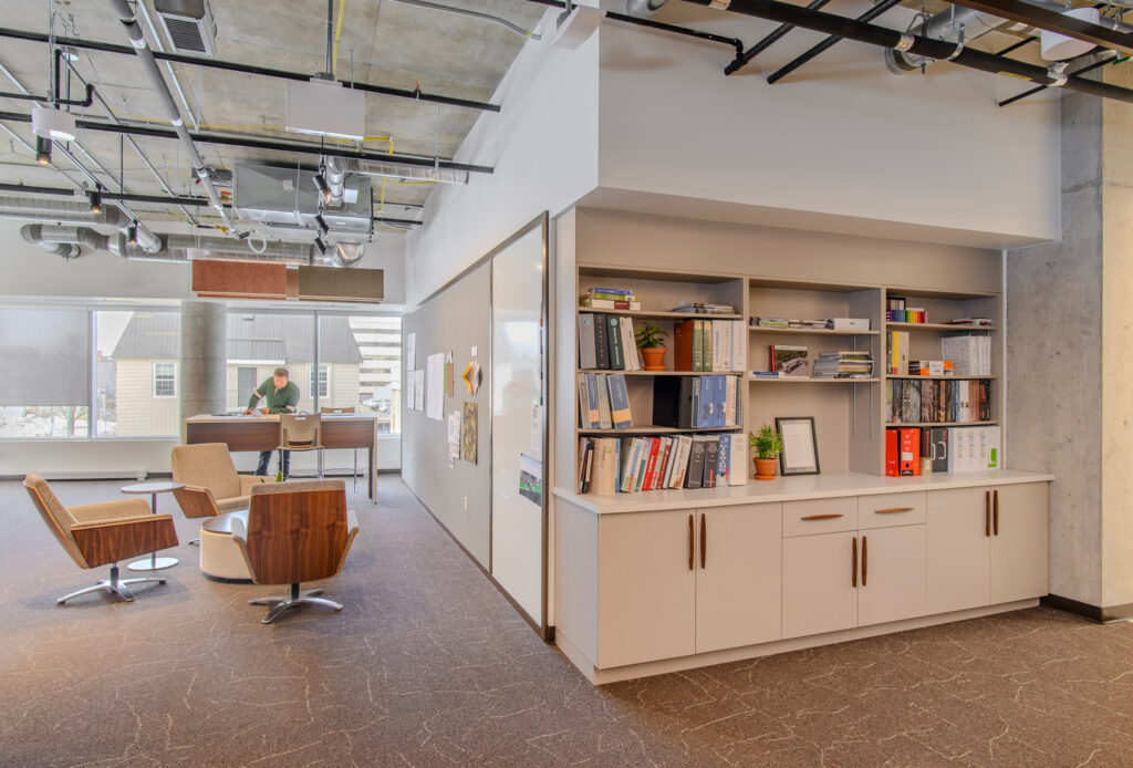 Modern office space featuring stylish interior design with open shelves for books and folders, a comfortable seating area with chairs, and a person working at a desk by large windows.