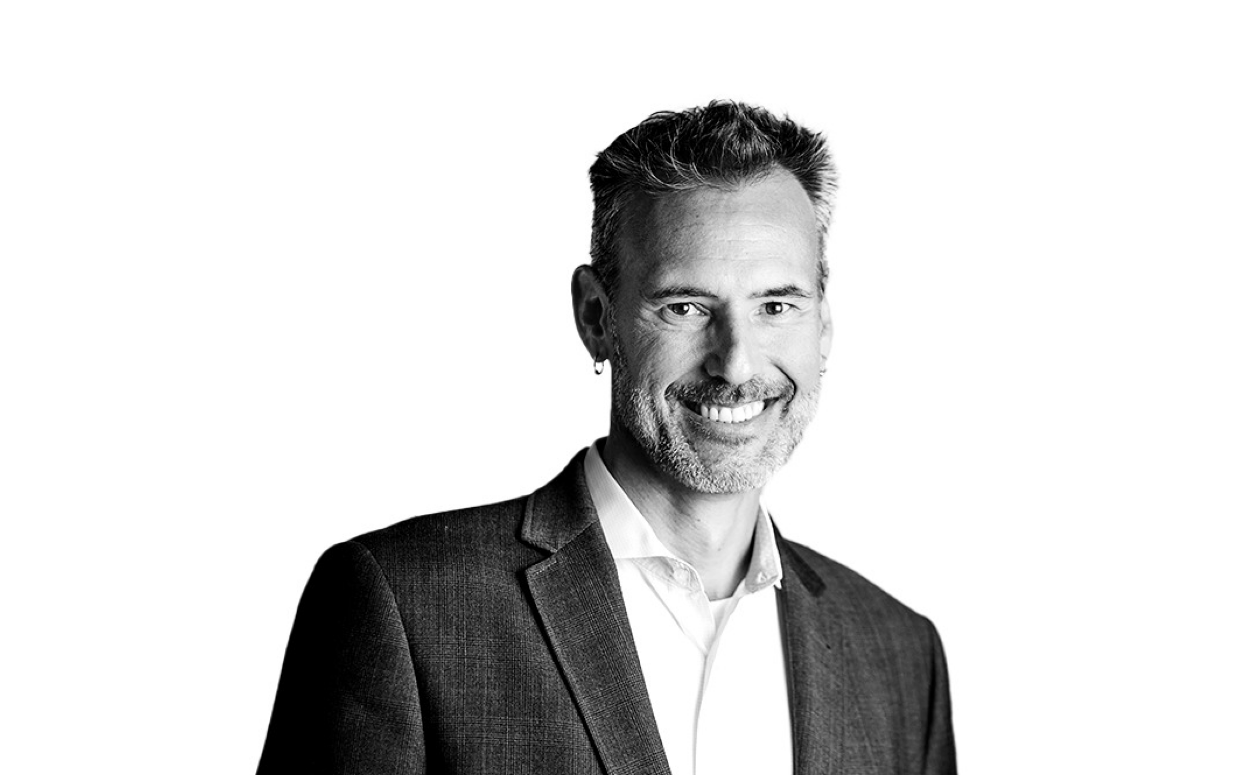 Portrait en noir et blanc d'un homme d'âge moyen aux cheveux courts et à la barbe, portant une veste de costume et une chemise blanche, souriant à l'appareil photo sur un fond uni.