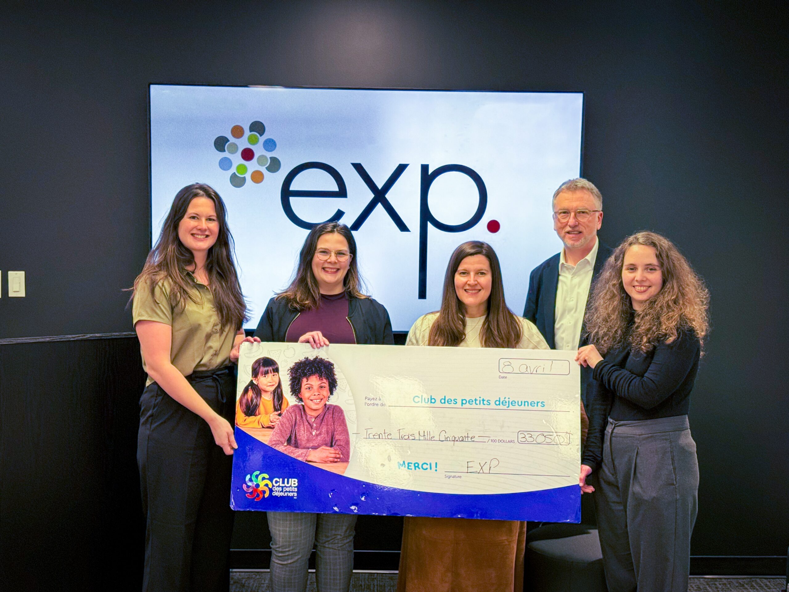 Cinq personnes se tiennent à l'intérieur et tiennent un gros chèque "Club des petits déjeuners" devant un écran affichant le logo "exp.