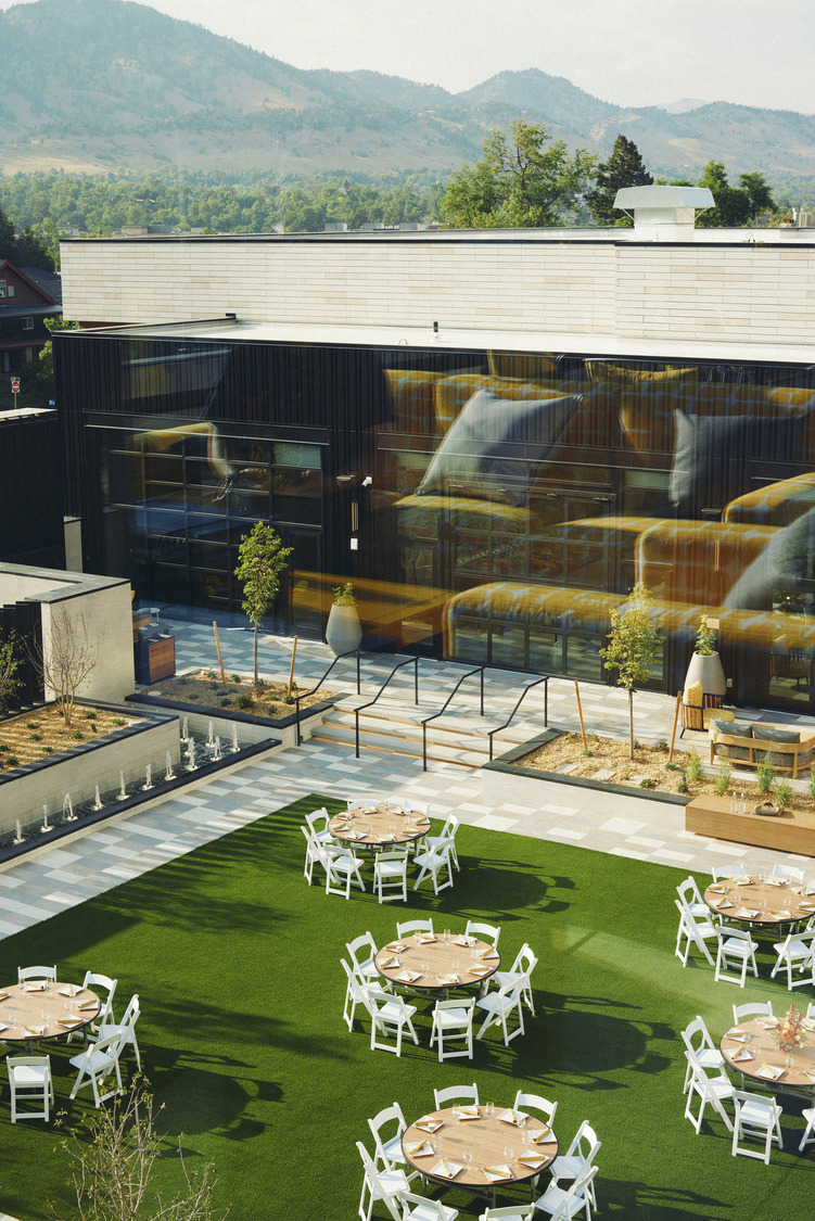 Round tables with white chairs are arranged on a green lawn outside the all-electric Limelight Boulder, a modern building with large windows and mountain views in the background.