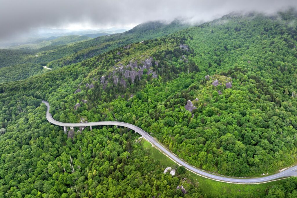 A winding road curves through a lush green forested mountain landscape under a cloudy sky.