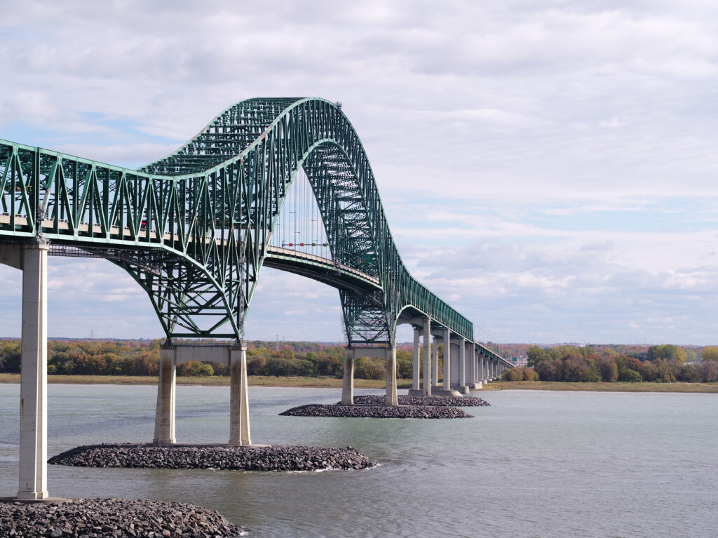 Un grand pont vert en arc d'acier enjambe une large rivière, soutenu par plusieurs piliers en béton, avec des arbres et un ciel nuageux en arrière-plan.