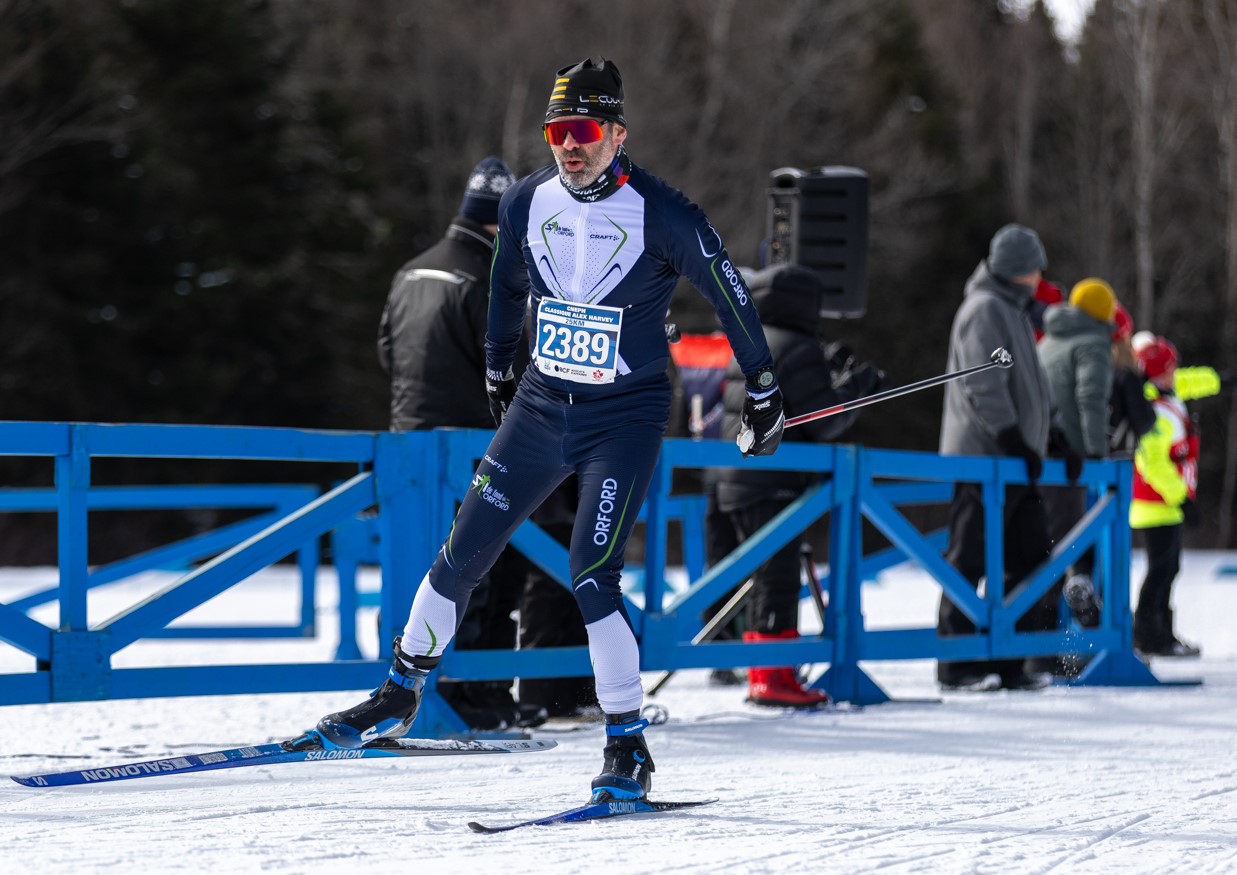 Un skieur de fond portant le numéro de dossard 2389 s'élance sur un parcours enneigé, sous le regard de spectateurs et d'officiels à l'arrière-plan.