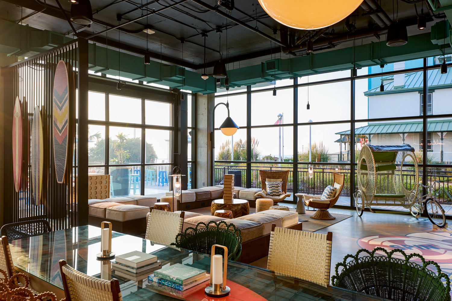 Modern lounge area with large windows, contemporary furniture, a mix of wooden and wicker chairs, and outdoor views with greenery and a building visible in the background.