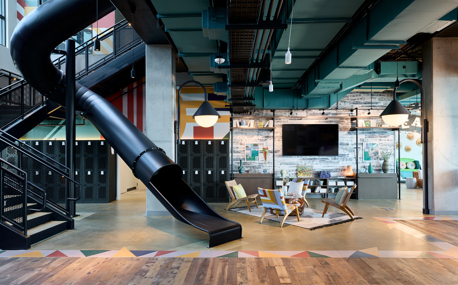 Modern office interior with a black slide, lockers, a seating area with chairs and a coffee table, industrial ceilings, and a staircase on the left.