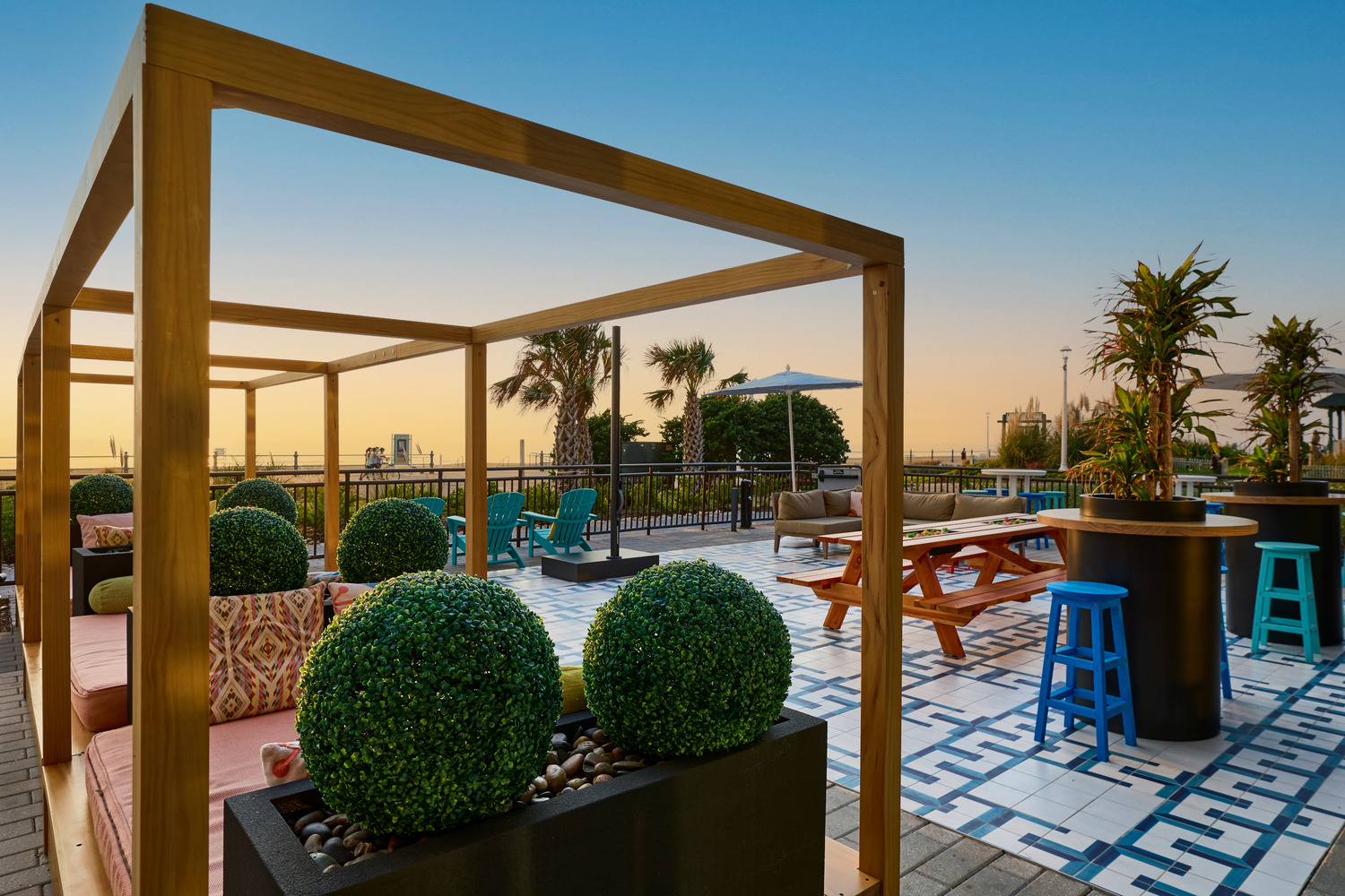 Modern rooftop patio at sunset with cabana seating, geometric tile flooring, potted plants, picnic tables, and blue stools; outdoor tables are arranged for casual gatherings.