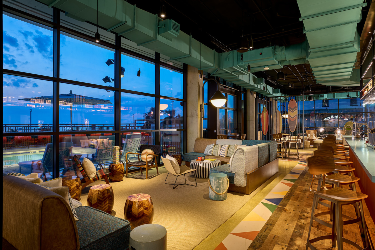 Modern lounge with eclectic furniture, bar stools, large windows, and city views at dusk; warm lighting creates a cozy atmosphere.