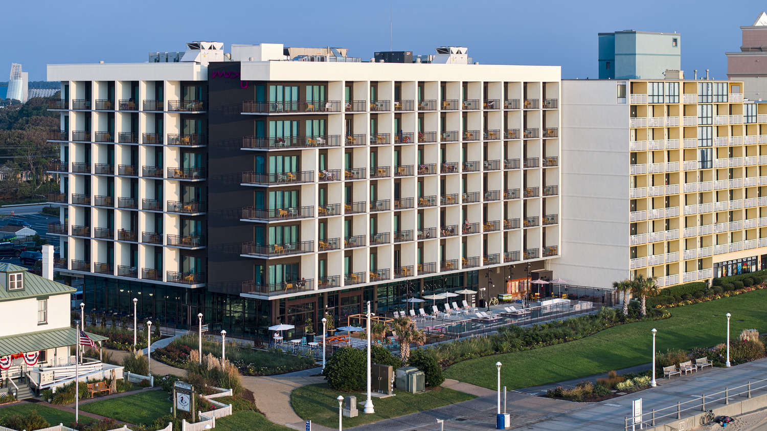 A modern, multi-story hotel with glass balconies overlooks a green lawn and pool area, adjacent to another hotel building in a coastal city setting.