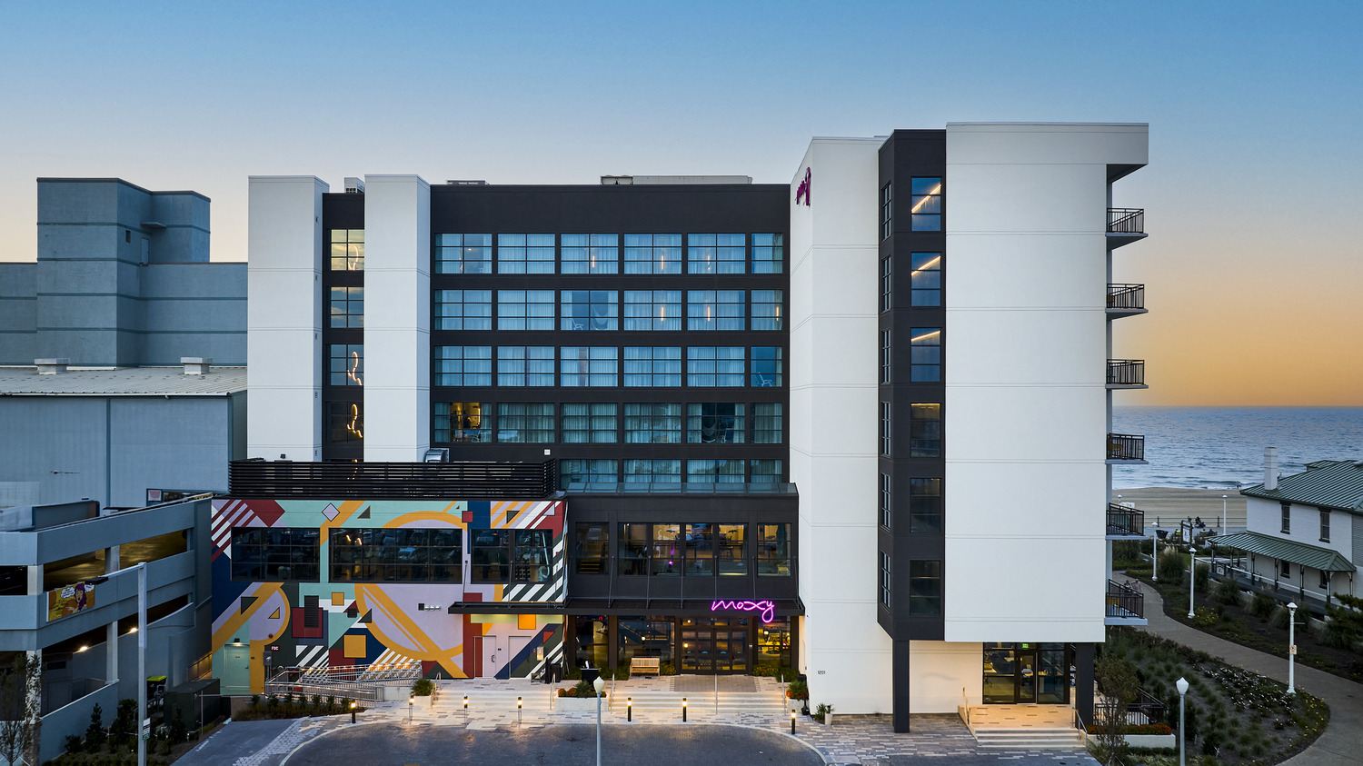 A modern beachfront hotel with colorful mural accents, large glass windows, and balconies, set against a backdrop of the ocean and a clear sky at sunset.