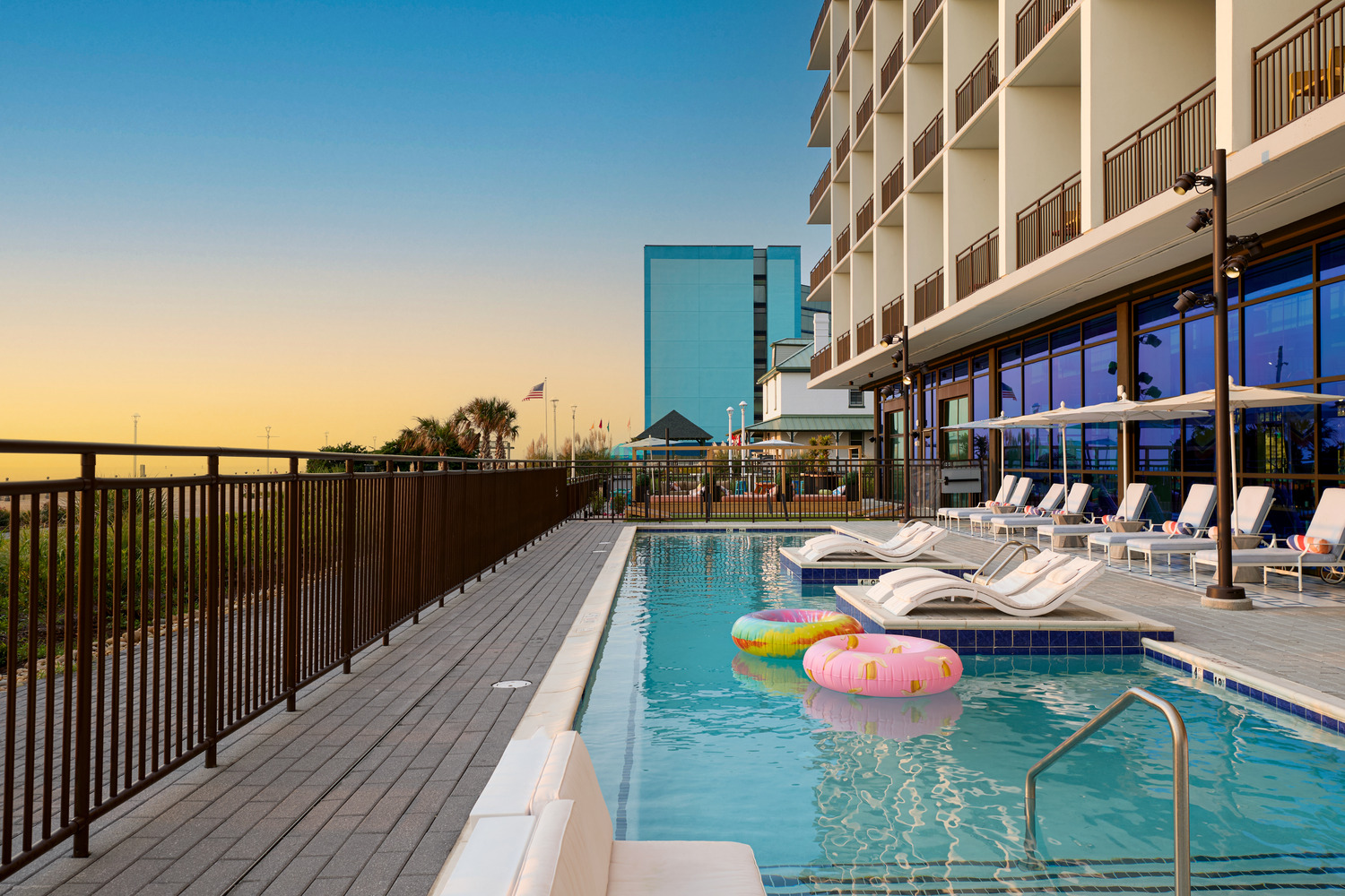 Outdoor hotel pool with lounge chairs, pool floats, and umbrellas alongside a modern building at sunset; a glass building and palm trees are visible in the background.