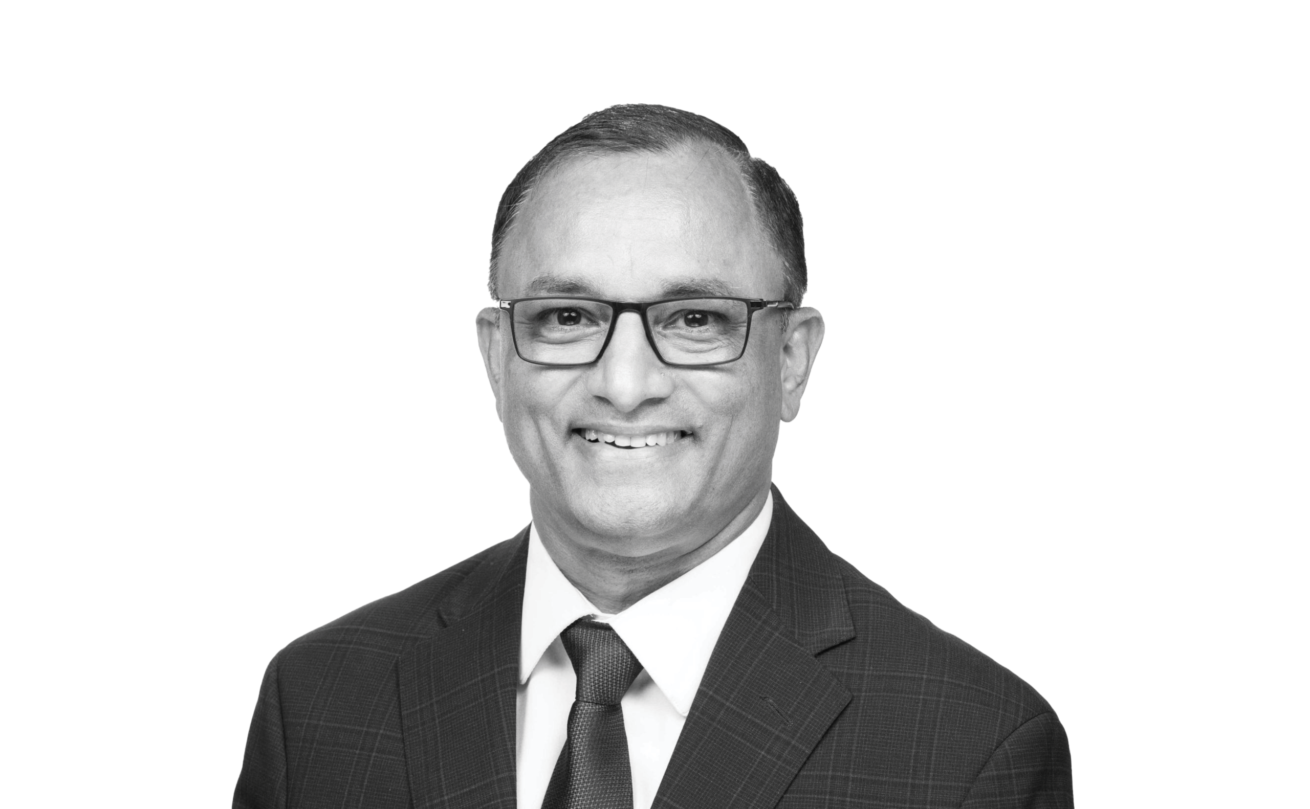 A man wearing glasses, a suit, and a tie smiles at the camera against a plain light background.