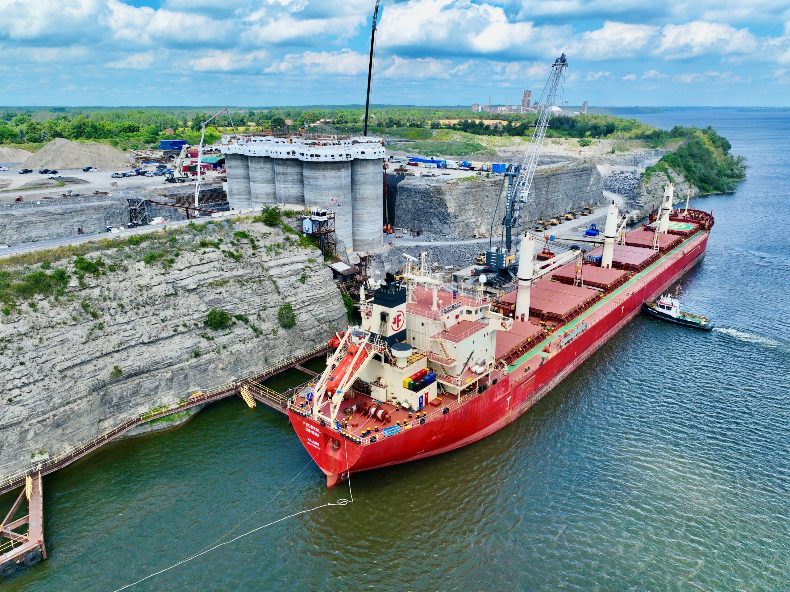 Un grand cargo rouge accosté dans un port industriel avec des grues et un remorqueur à proximité, entouré de rochers et d'eau sous un ciel partiellement nuageux.