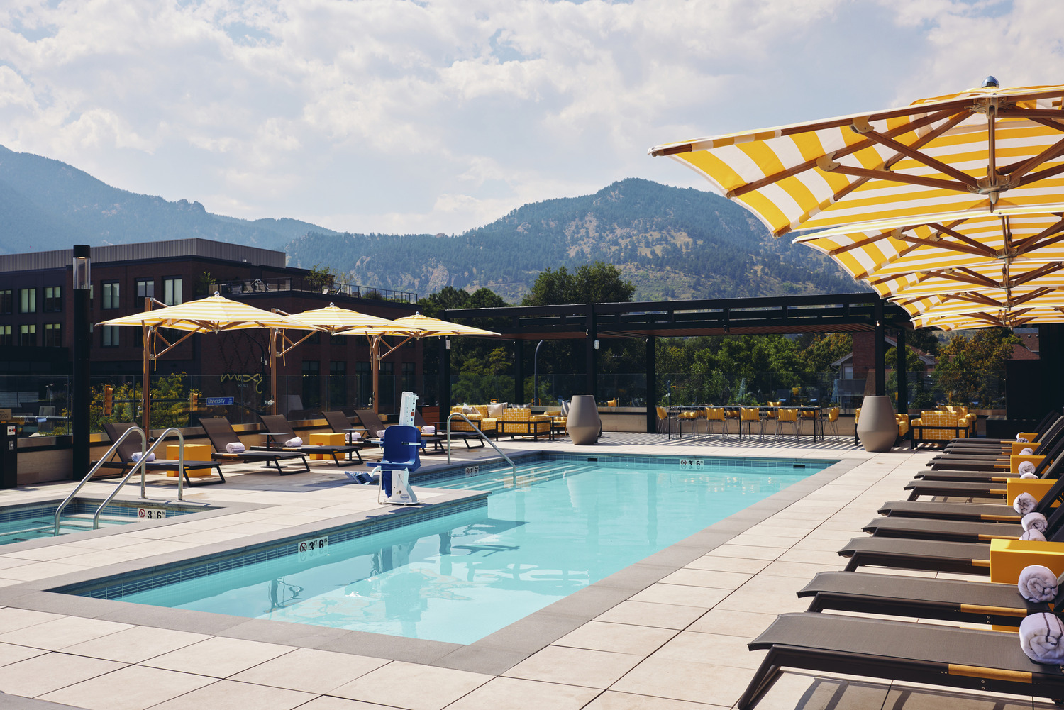 All-electric outdoor pool area with lounge chairs, striped yellow umbrellas, and mountain views in the background at Limelight Boulder on a partly cloudy day.