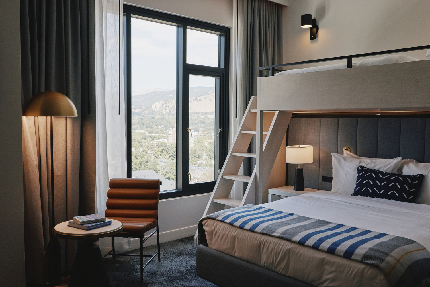 A modern, all-electric hotel room at Limelight Boulder features a bunk bed, double bed, mountain-view window, lounge chair, floor lamp, and a small round table with books.