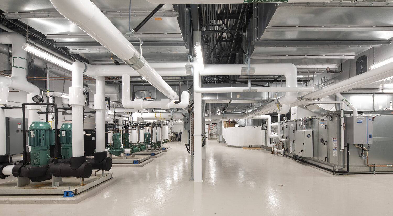 A clean, brightly lit industrial mechanical room with large pipes, green pumps, and HVAC equipment lining the walls and ceiling.