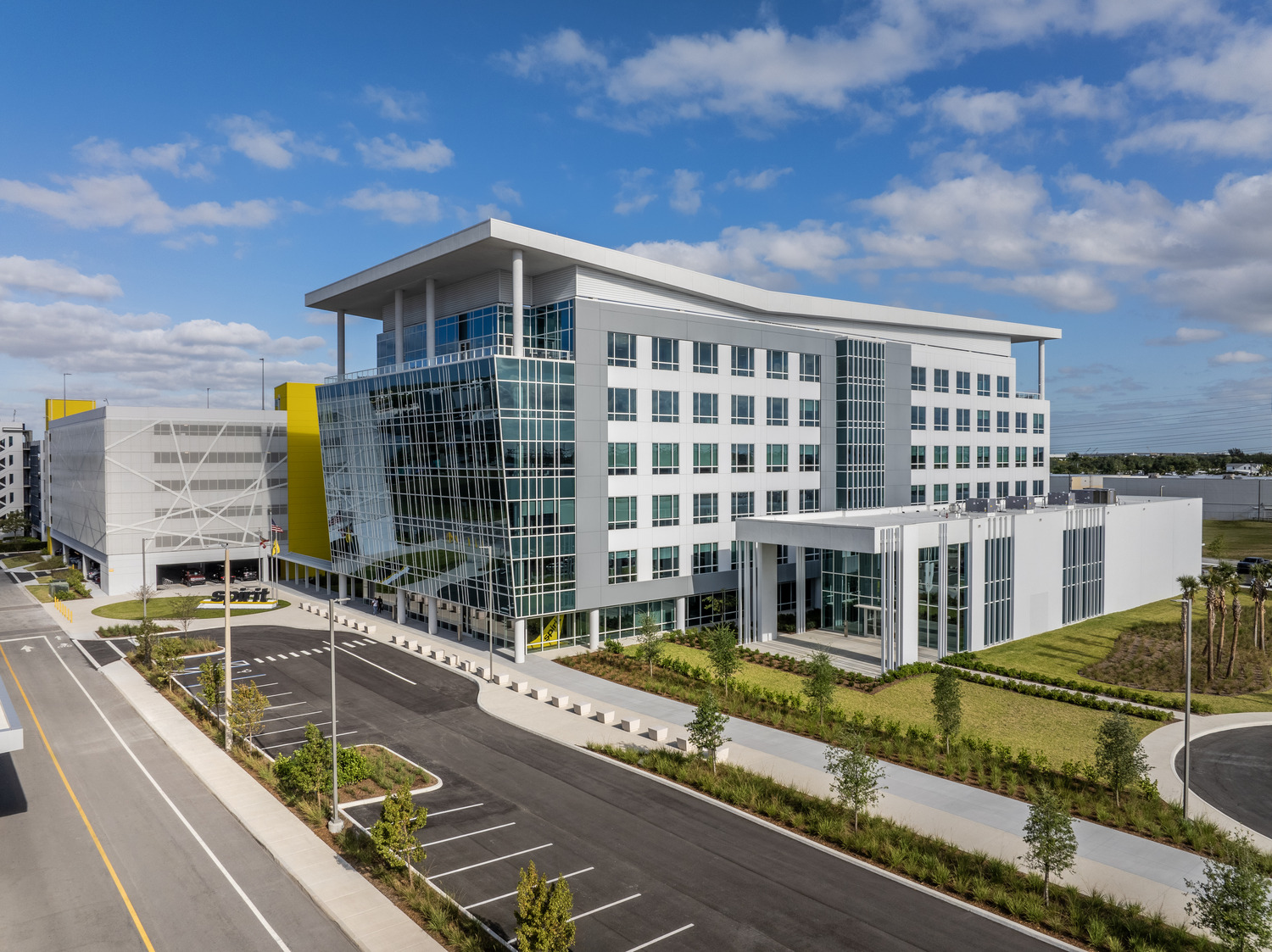 A modern multi-story office building with large windows and white exterior, surrounded by landscaped grounds and adjacent to a parking structure under a blue sky with scattered clouds.