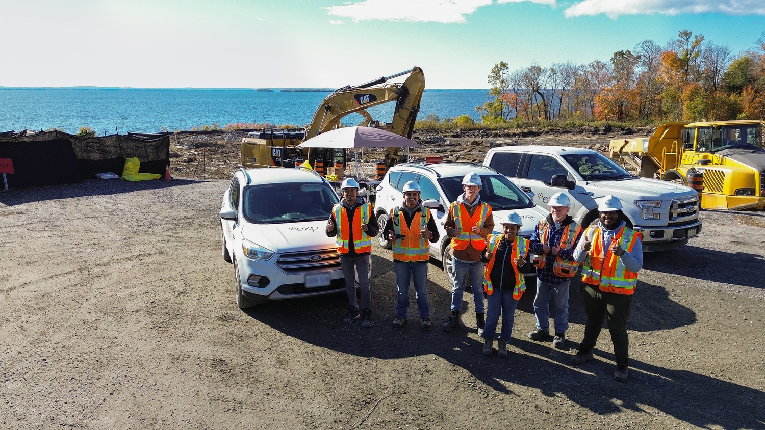 Un groupe de travailleurs de la construction portant des gilets de sécurité et des casques se tient devant des véhicules et des équipements sur un chantier situé à proximité d'un plan d'eau.