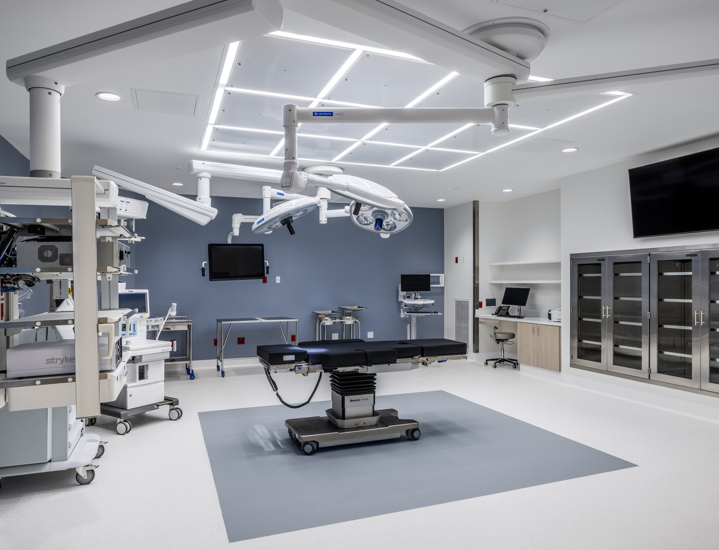 Modern operating room with surgical lights, an operating table, medical equipment, monitors, storage cabinets, and a computer workstation. Room is clean, well-lit, and unoccupied.