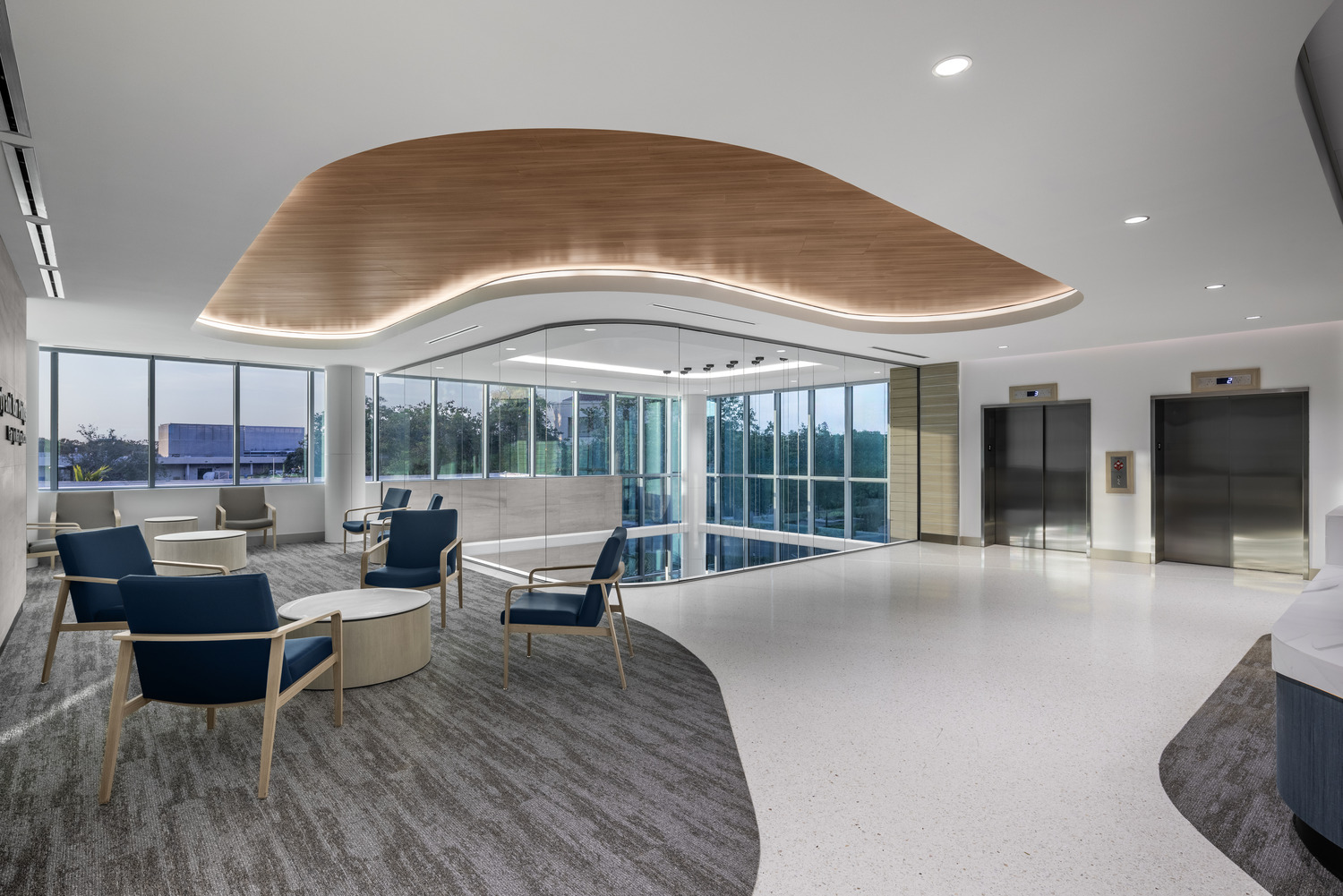 Modern hospital waiting area with blue chairs, round tables, large windows, two elevators, and a curved wooden ceiling accent.
