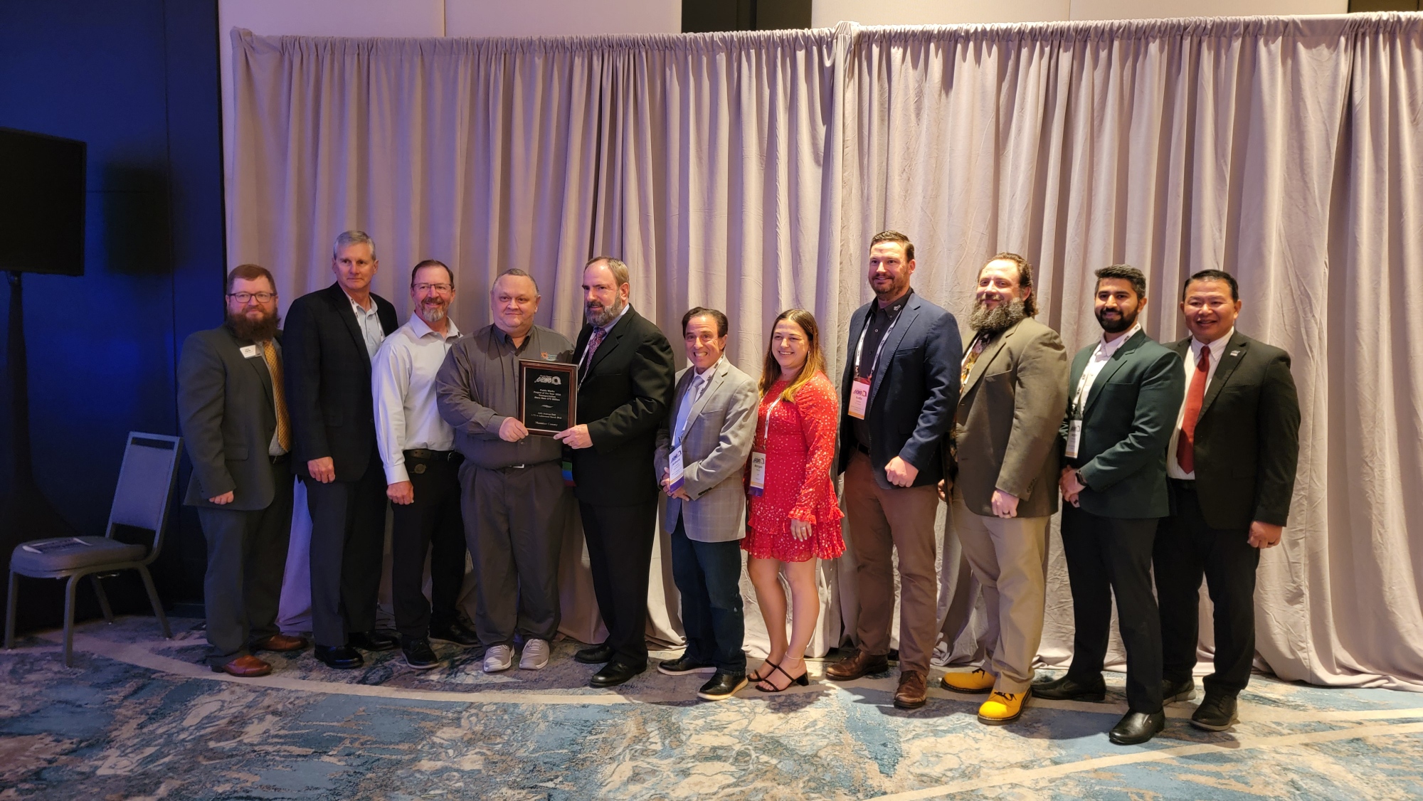 A group of eleven people stands in a row indoors; one person in the center holds a plaque recognizing the 44th Avenue East Extension, honored by APWA. They are dressed in business or business casual attire.