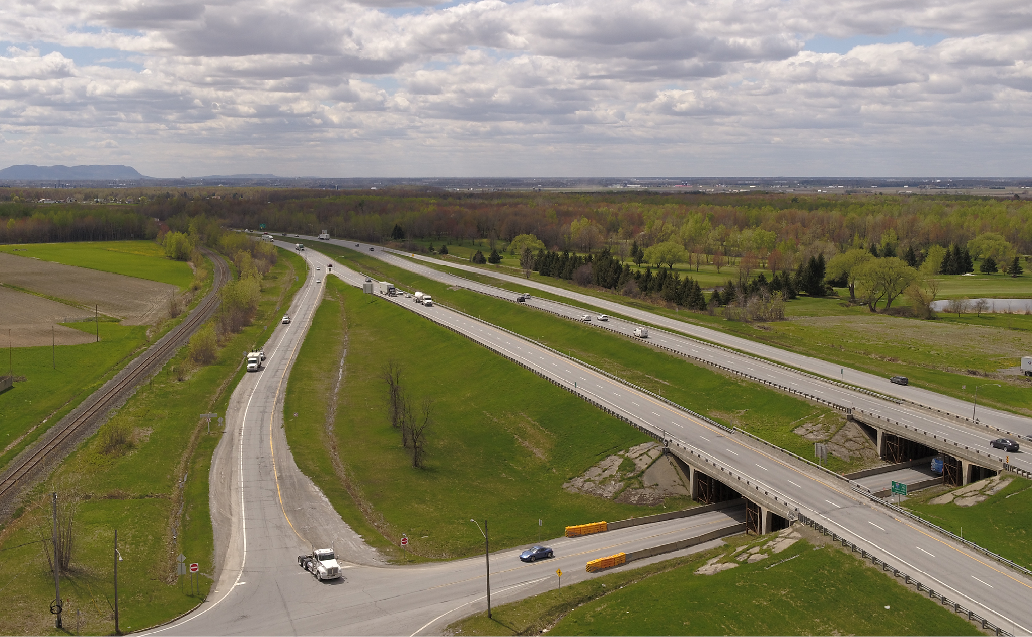 Aerial view of a highway interchange with several cars and trucks, surrounded by green fields, trees, and cloudy sky in the background.
