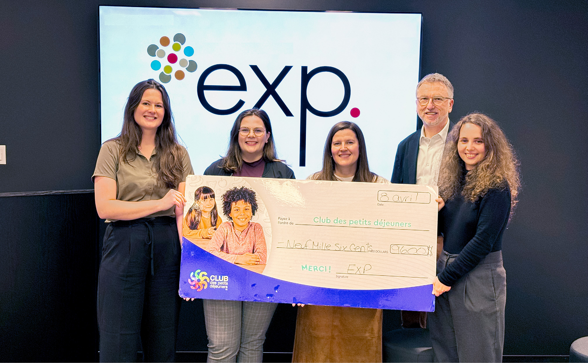 Cinq personnes se tiennent à l'intérieur, tenant un chèque surdimensionné de 1 600 dollars à l'ordre du Club des petits déjeuners, avec un logo "exp." affiché sur un écran derrière elles.