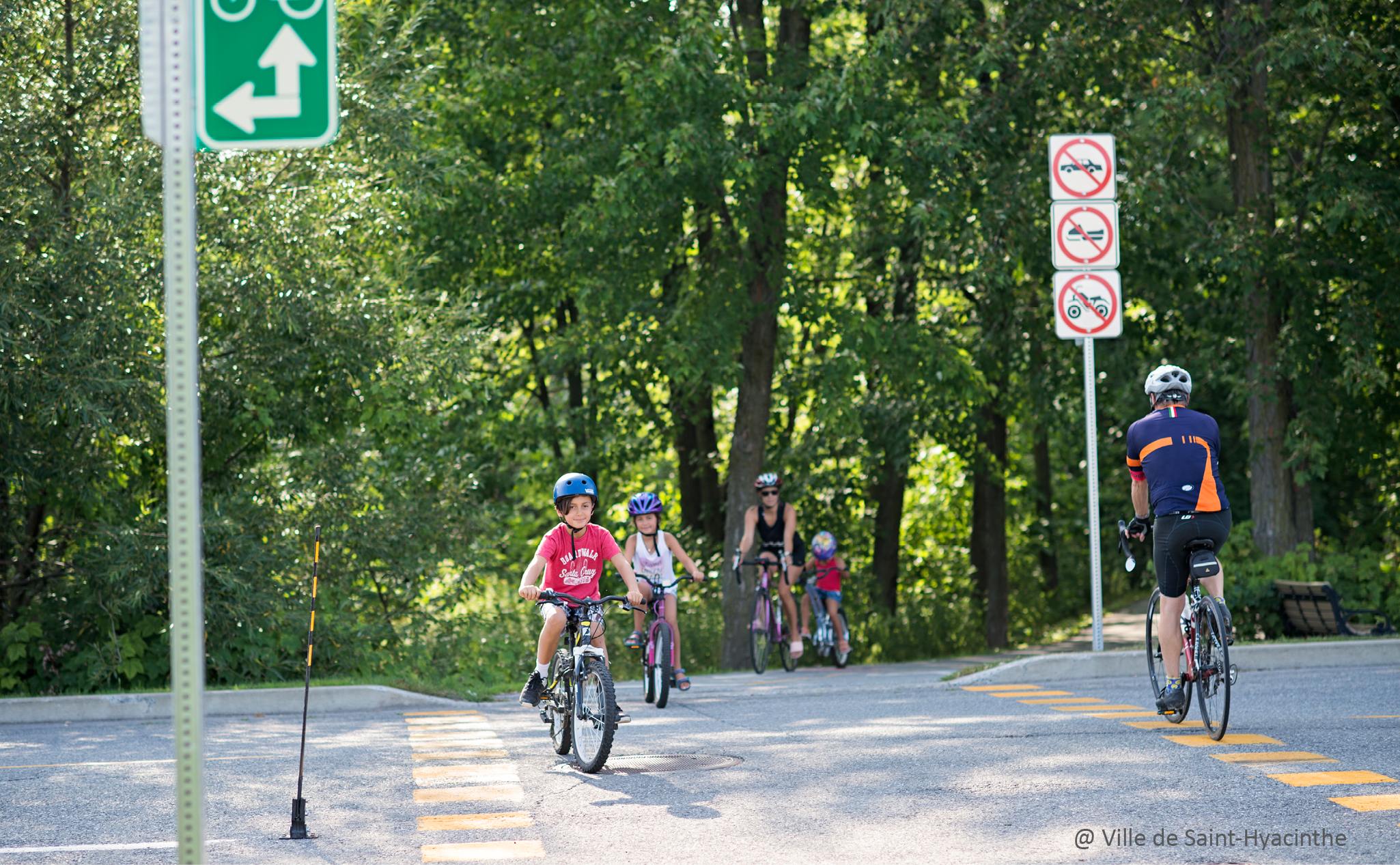 Plusieurs personnes, dont des enfants et des adultes, font du vélo sur un sentier dans un parc boisé, à proximité de panneaux d'interdiction d'accès et de chiens.