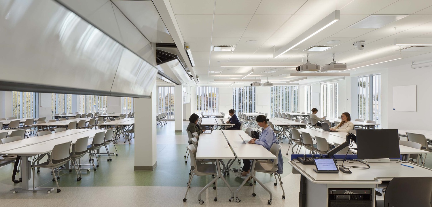 Modern classroom with large windows, several students seated at tables, and a computer workstation in the foreground. The room is bright with natural light.