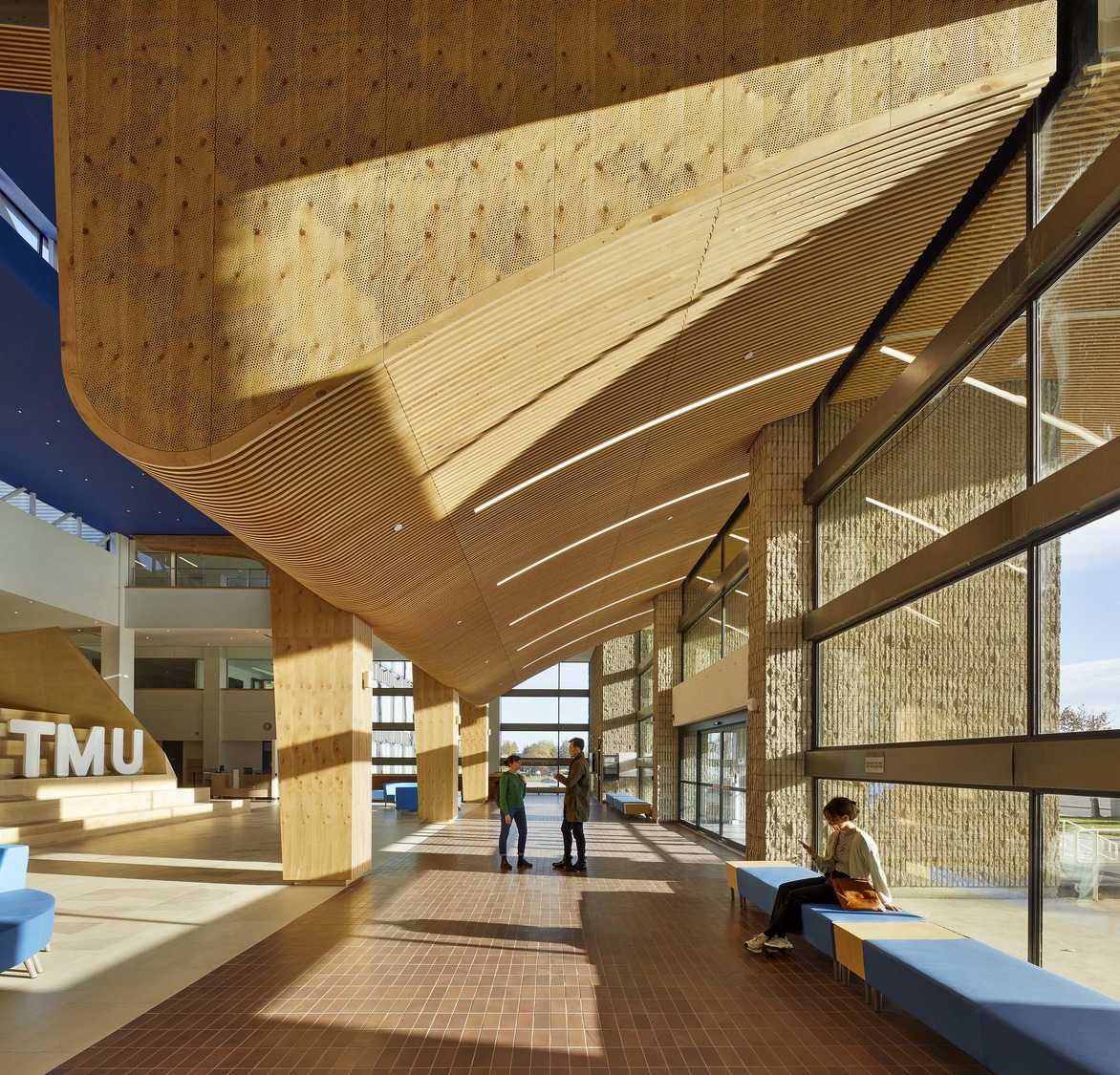 Spacious modern lobby with wood paneling, large windows, blue seating, and three people walking while one person sits near the entrance.