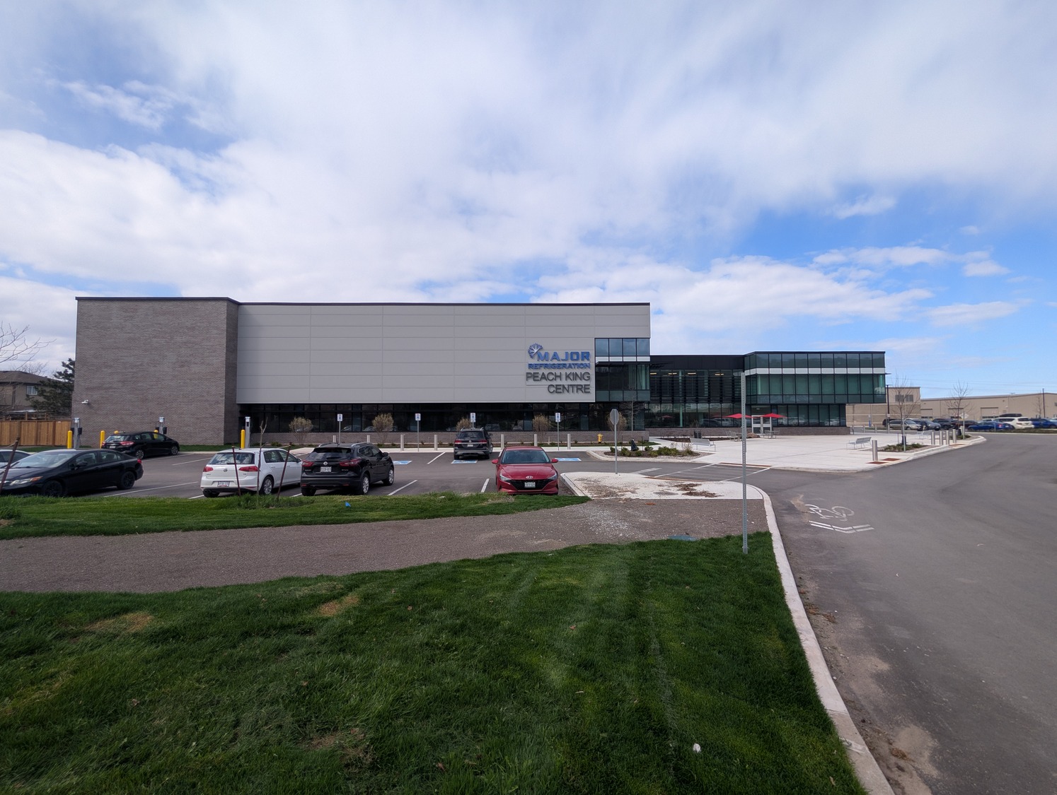 A modern building labeled "REALTOR REACHING CENTRE," located near the Peach King Centre, stands with a parking lot, several parked cars, and a grassy area in the foreground under a partly cloudy sky.