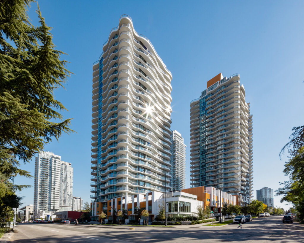 Two modern high-rise residential buildings with curved balconies stand along a city street on a clear, sunny day.